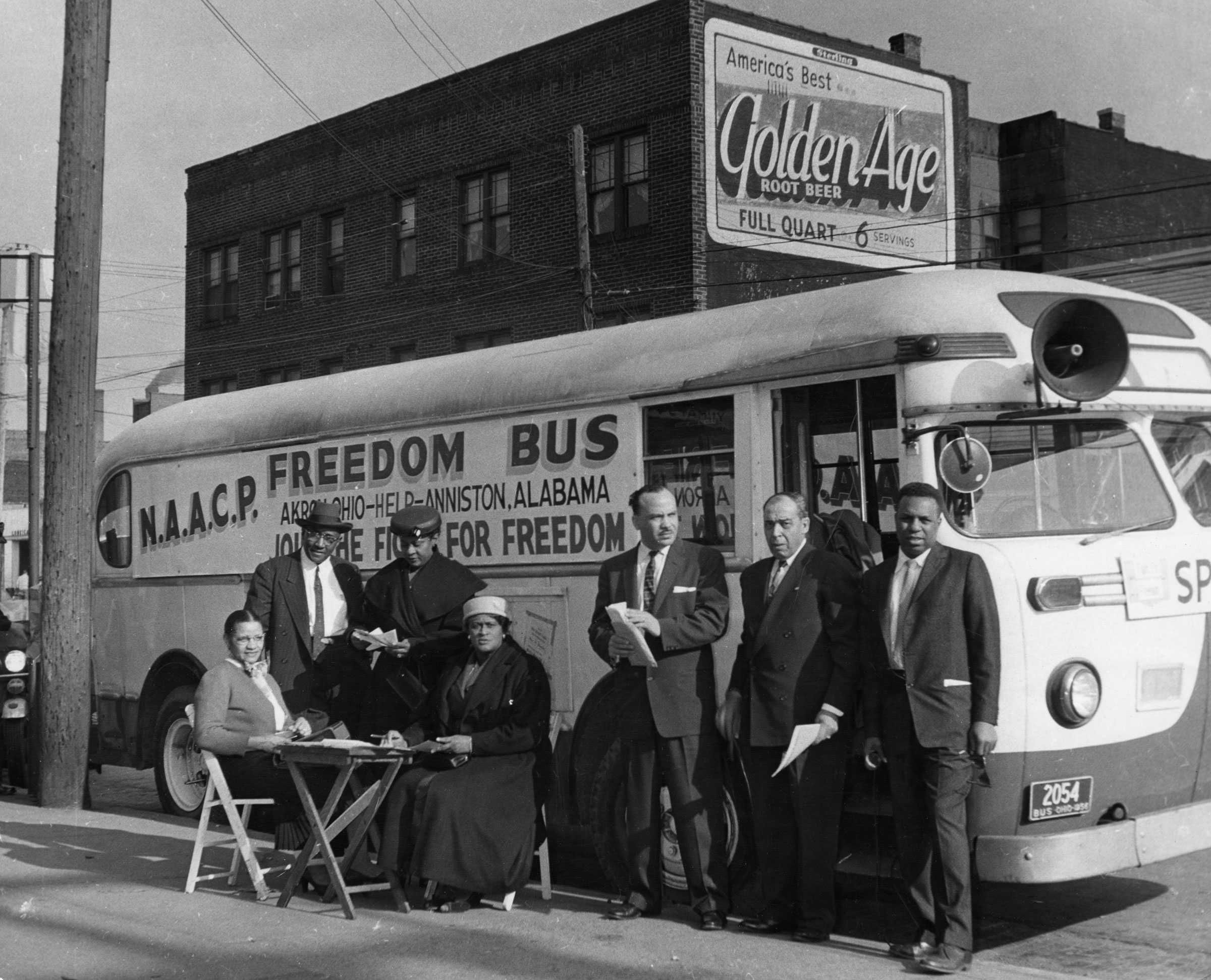 NAACP members gather outside of a Freedom Bus parked on a street. A few members are sitting down at a folding chair.