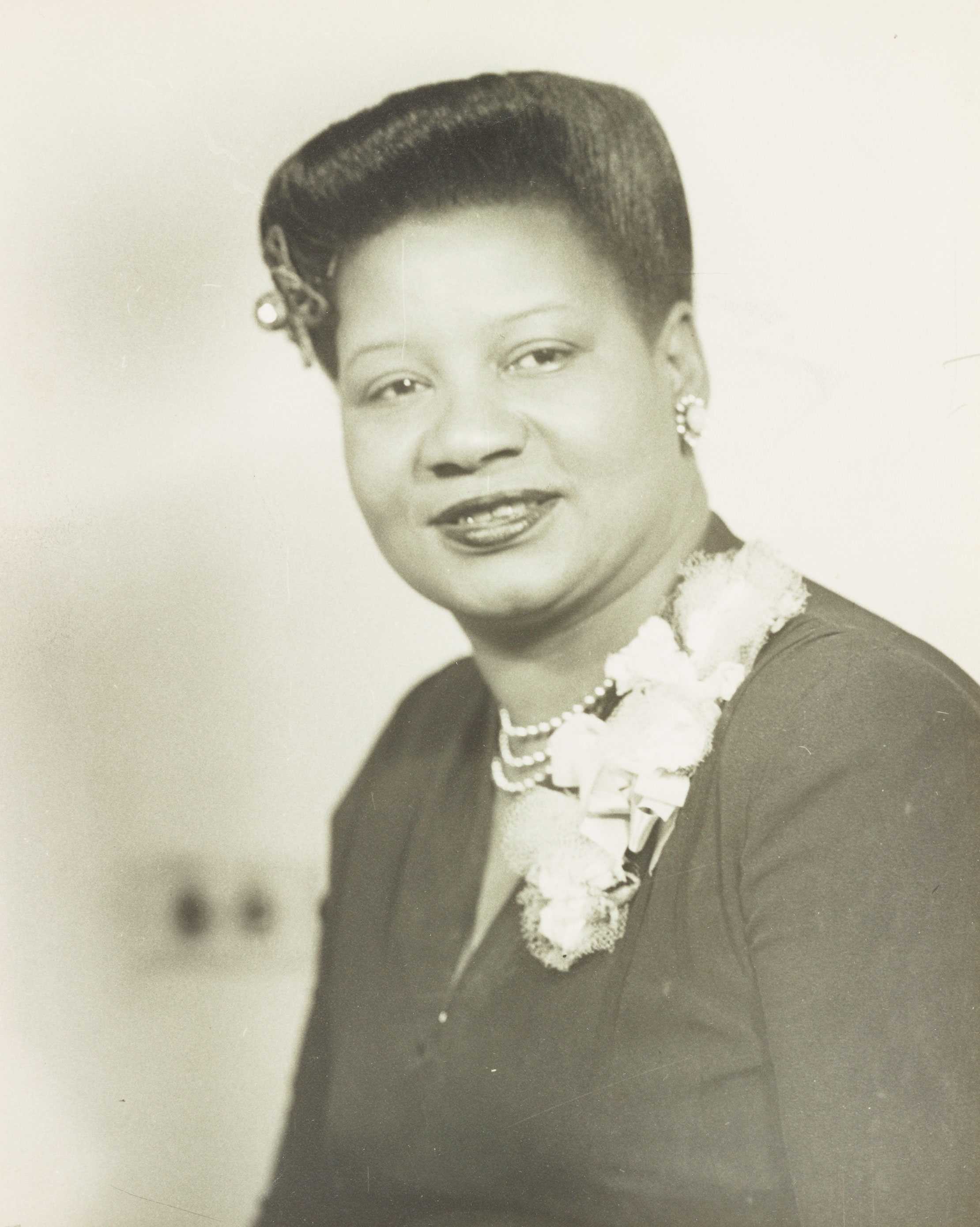 A black-and-white portrait of a woman with earrings, a pearl necklace, and bows on her left shoulder.