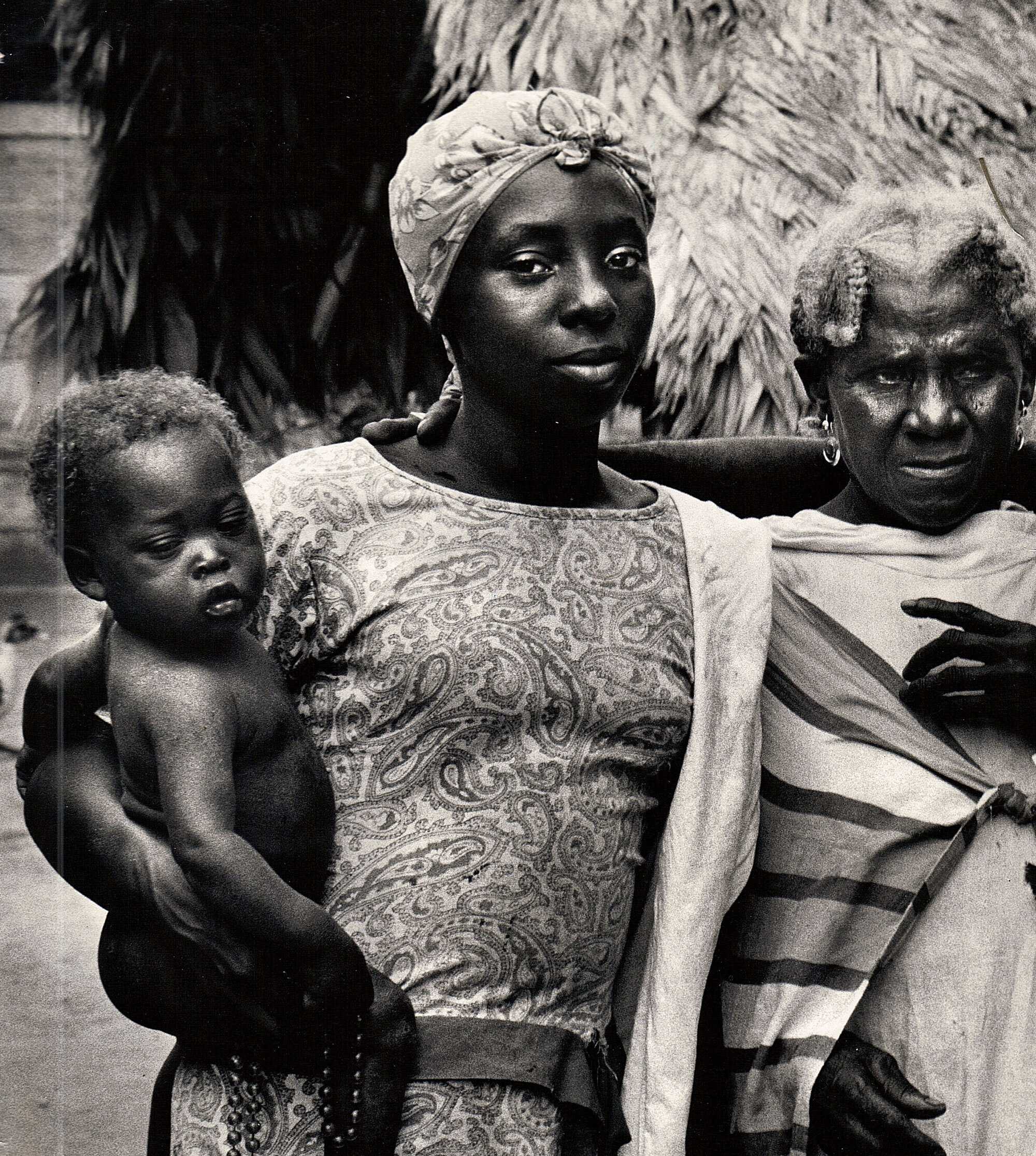 A black and white photograph ofa family. A mother is holding a child, while standing next to an elderly woman.