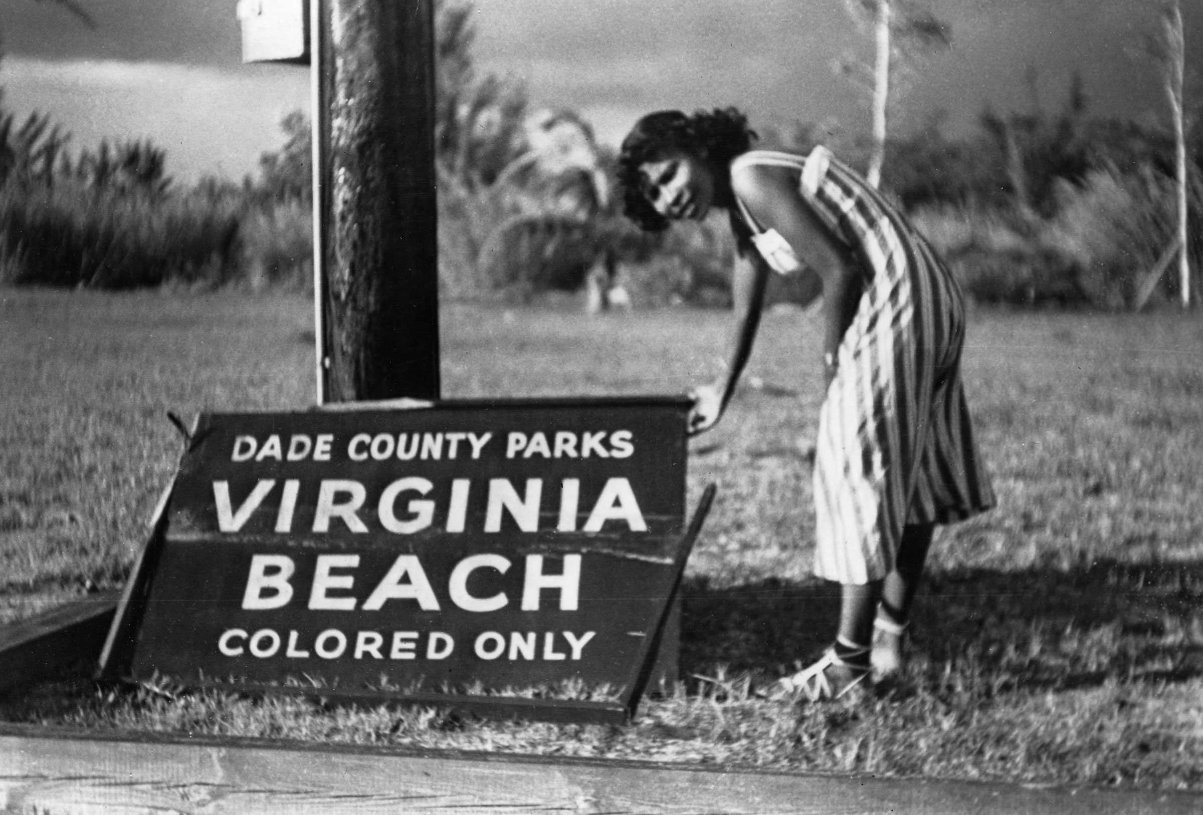 Black and white photograph showing an African American woman dressed in a striped sun dress holding a sign that has fallen to the ground.  The sign says "DADE COUNTY PARKS / VIRGINIA / BEACH / COLORED ONLY."