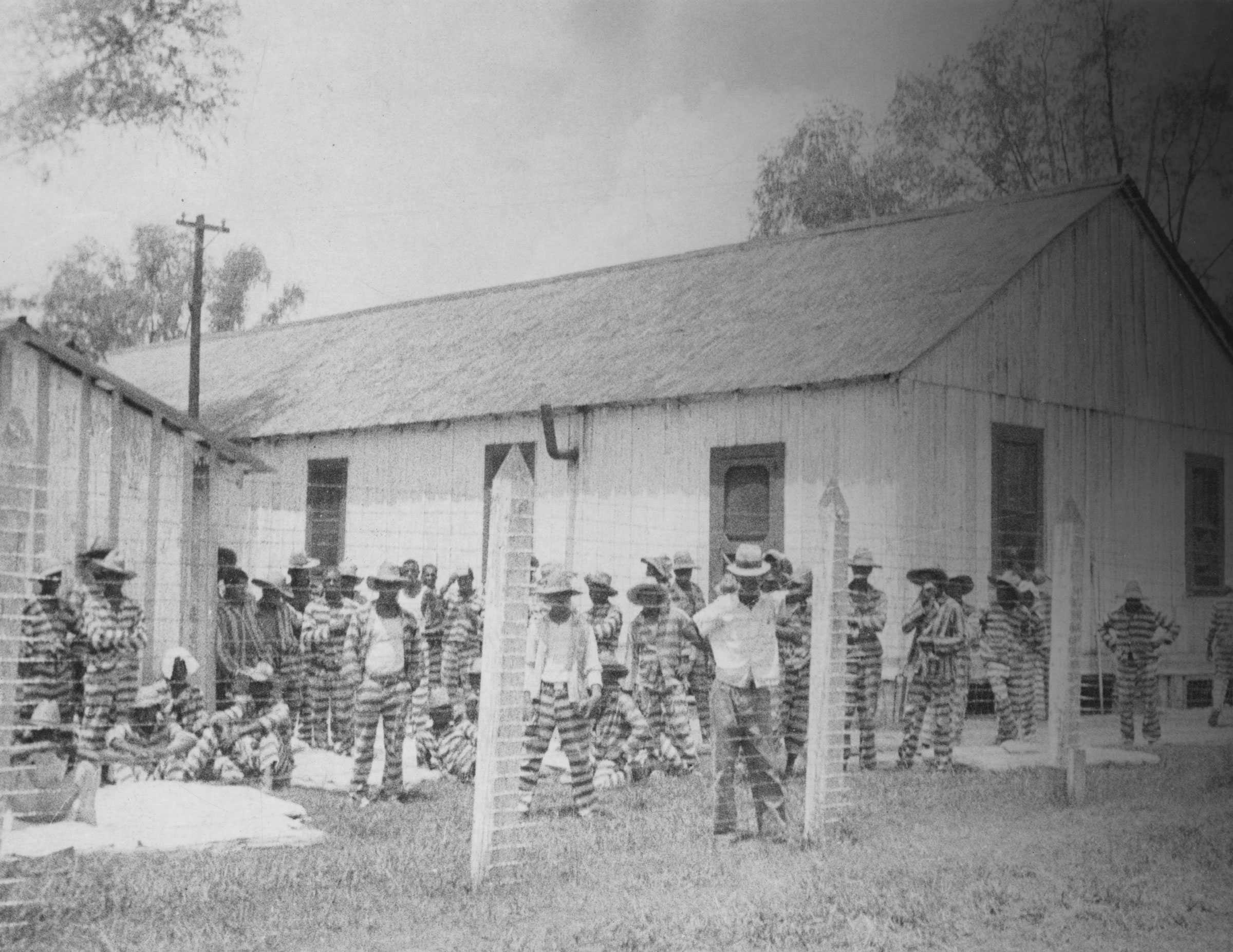 Black and white photograph of prisoners behind a wired fence with buildings in the background.  The men are all wearing striped pants and shirts.