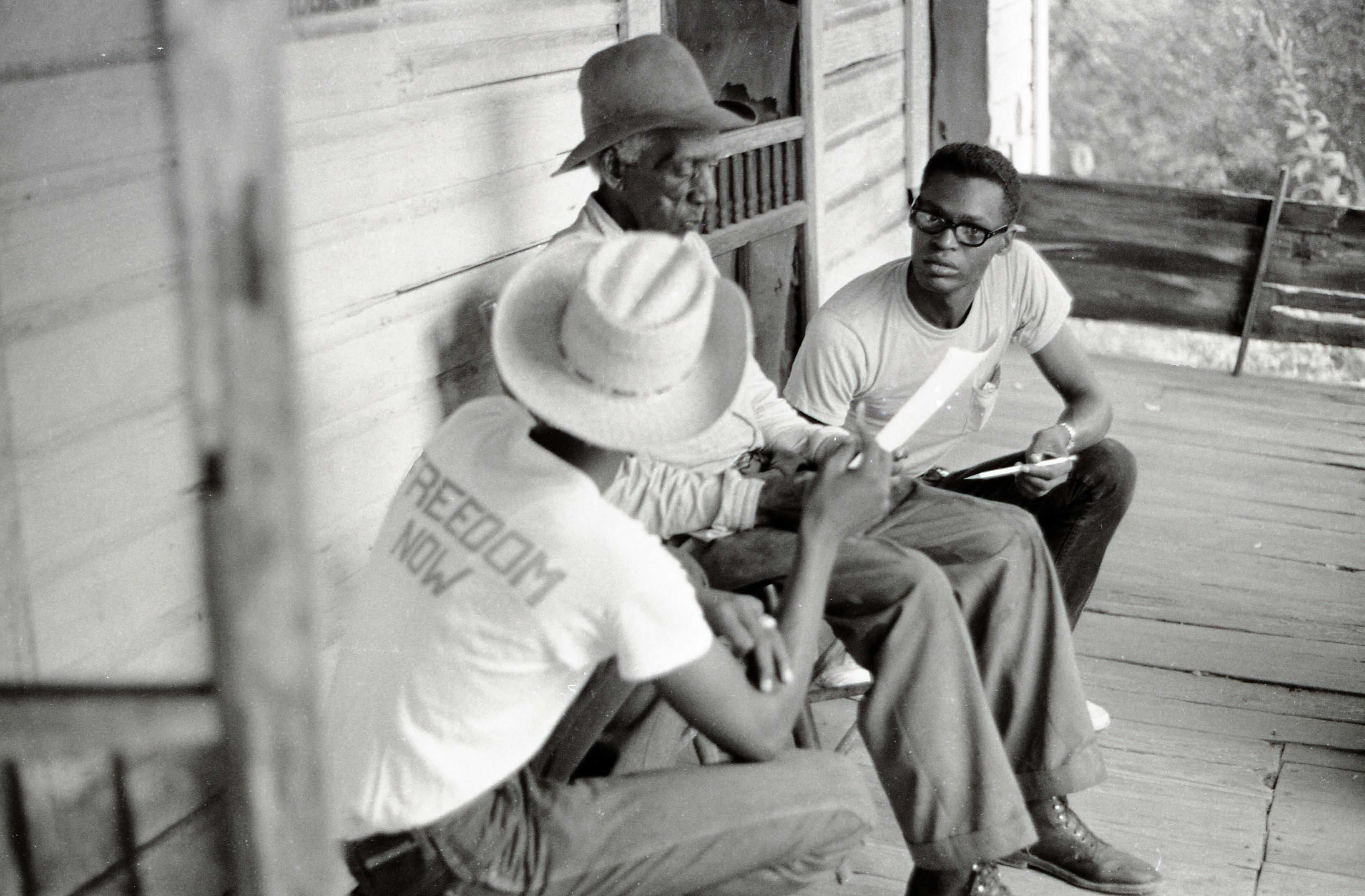 Black and white photograph showing three men sitting on a porch talking.