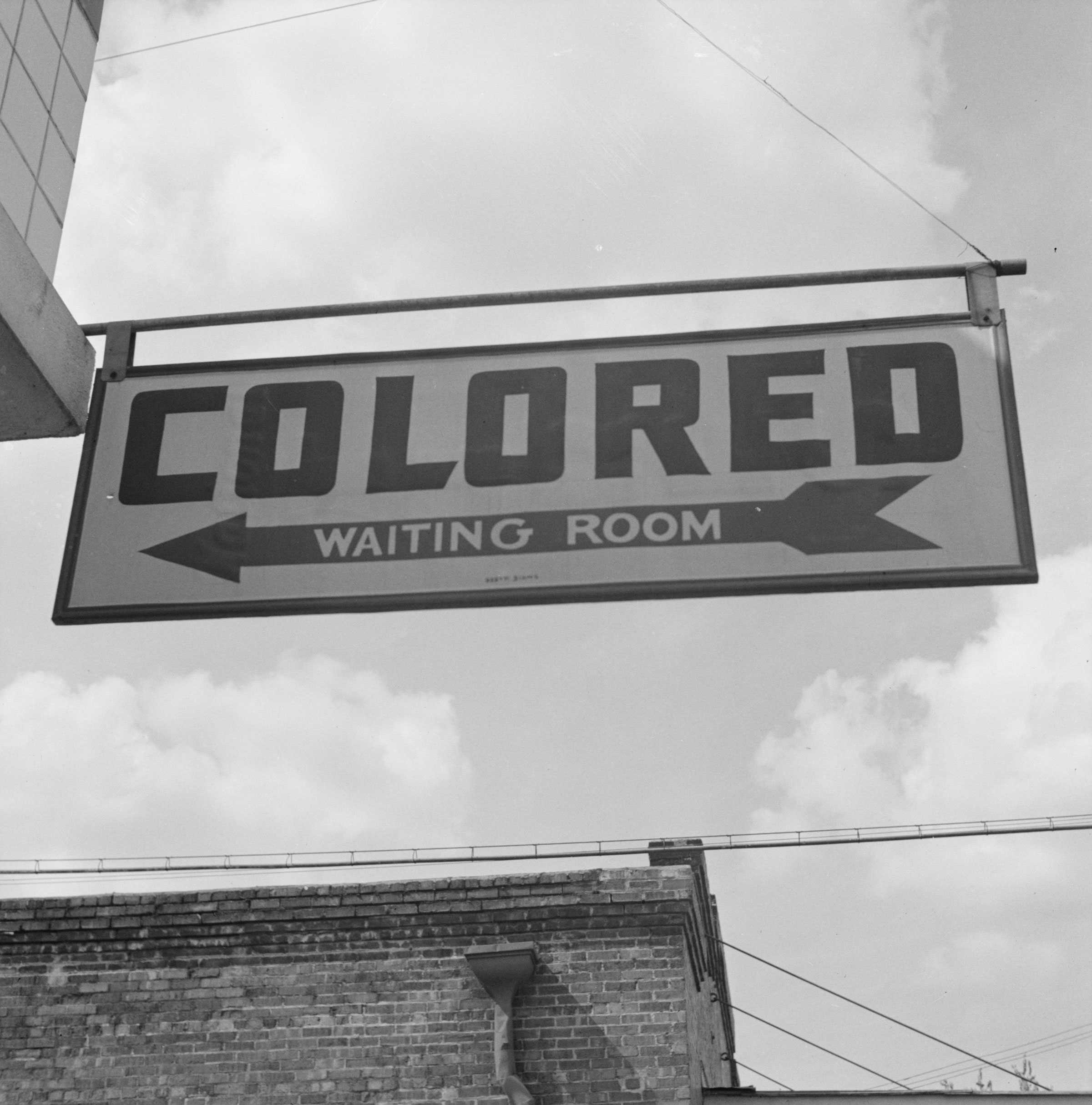 Black and white photograph of sign attached to building reading "COLORED / WAITING ROOM."