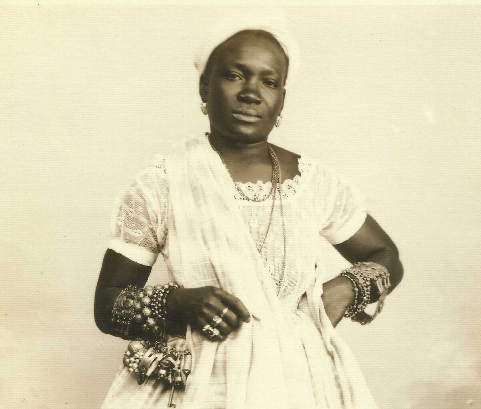 A sepia tone photgraph of a woman dressed in long white dress. She lots of chunky bangles on her arms.