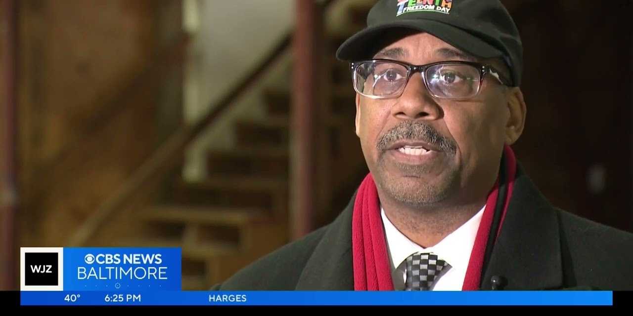 Screenshot from a news video showing an African American woman wearing glasses (Dr. Iris, the director of the Hosanna School Museum) seated at a wooden classroom desk.