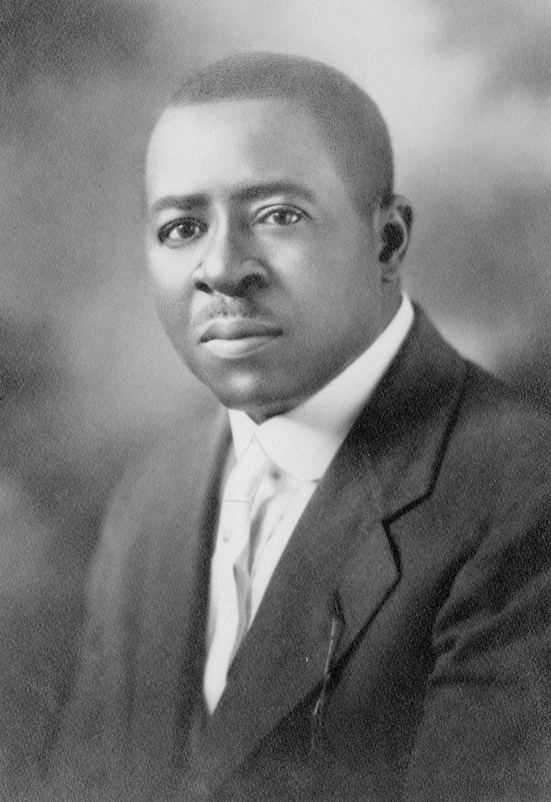 Portrait of a young Black man dressed in a suit coat and white shirt and tie.  The man gazes directly at the viewer.