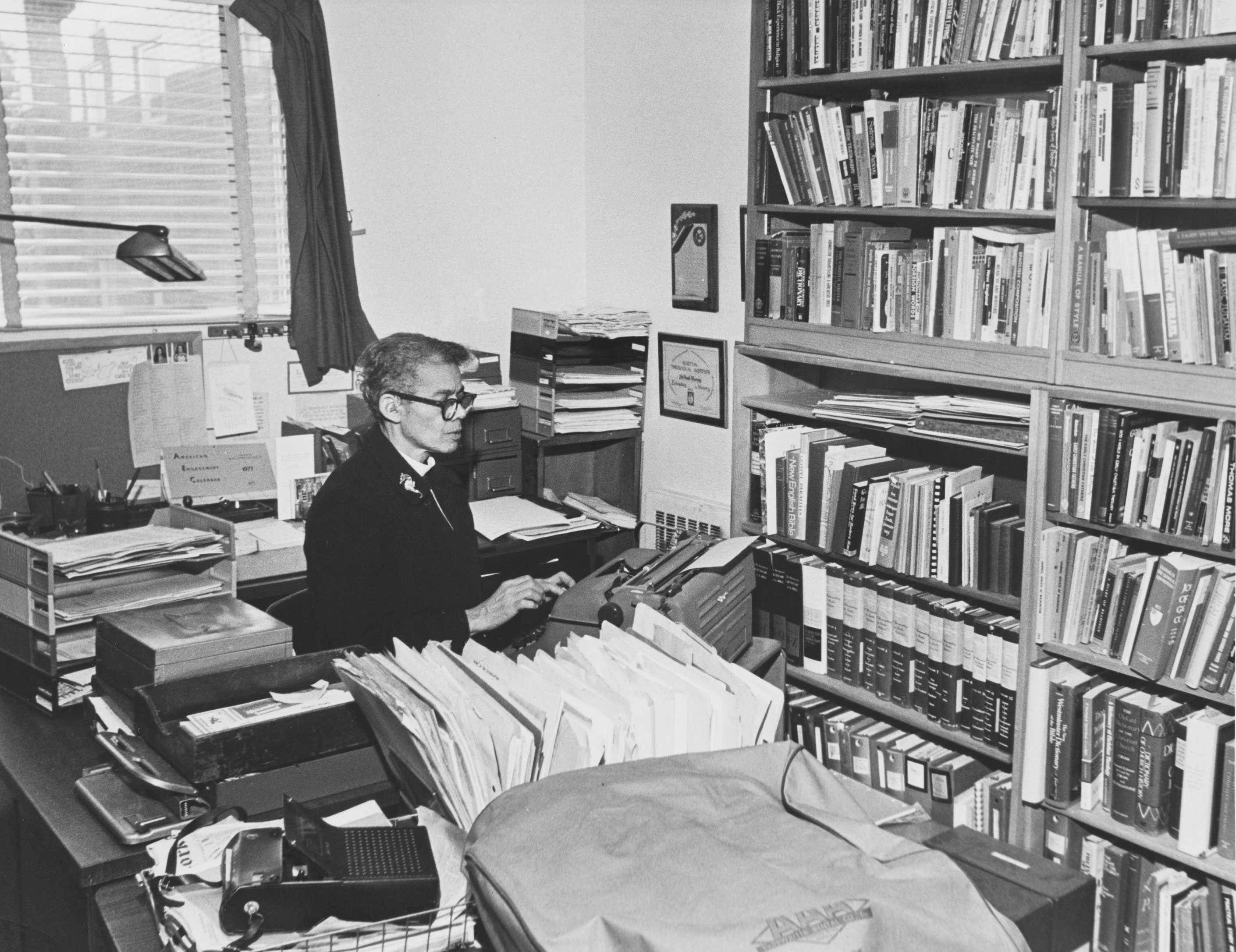 A black-and-white photograph of the Reverend Dr. Pauli Murray sitting at a typewriter by a desk crowded.