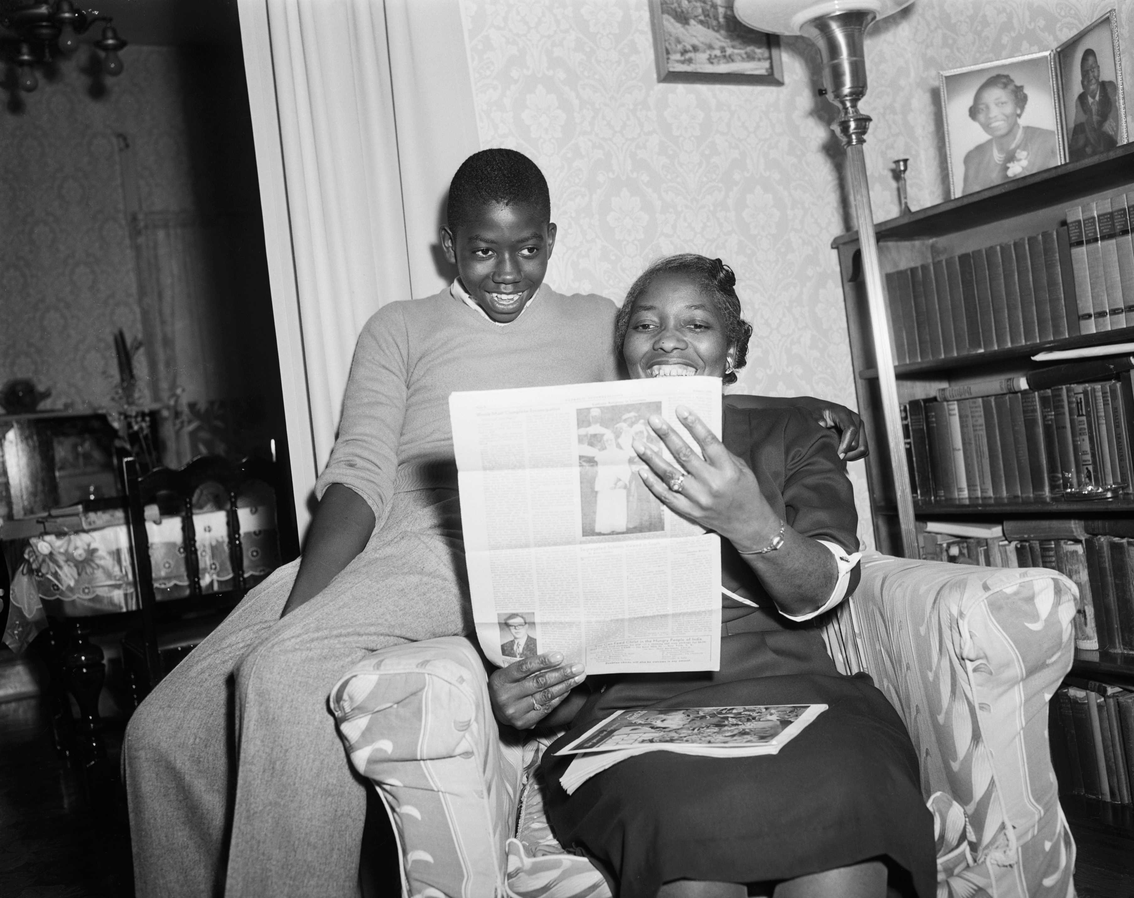 Black and white photograph of woman sitting in living room reading a newspaper to a boy seated on the arm of the chair.