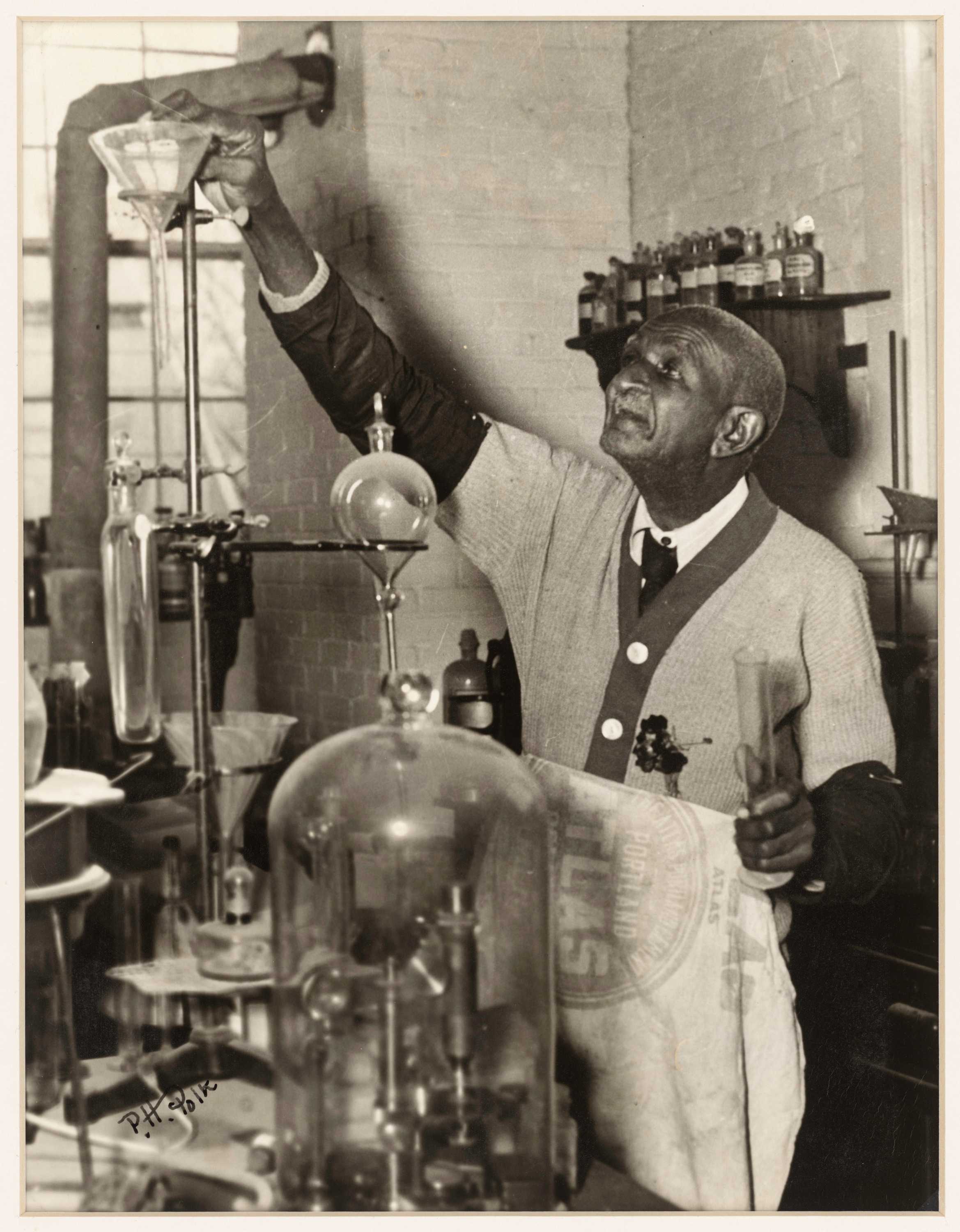 Black and white photograph of George Washington Carver shown in a laboratory wearing an apron and holding a beaker in his left hand.