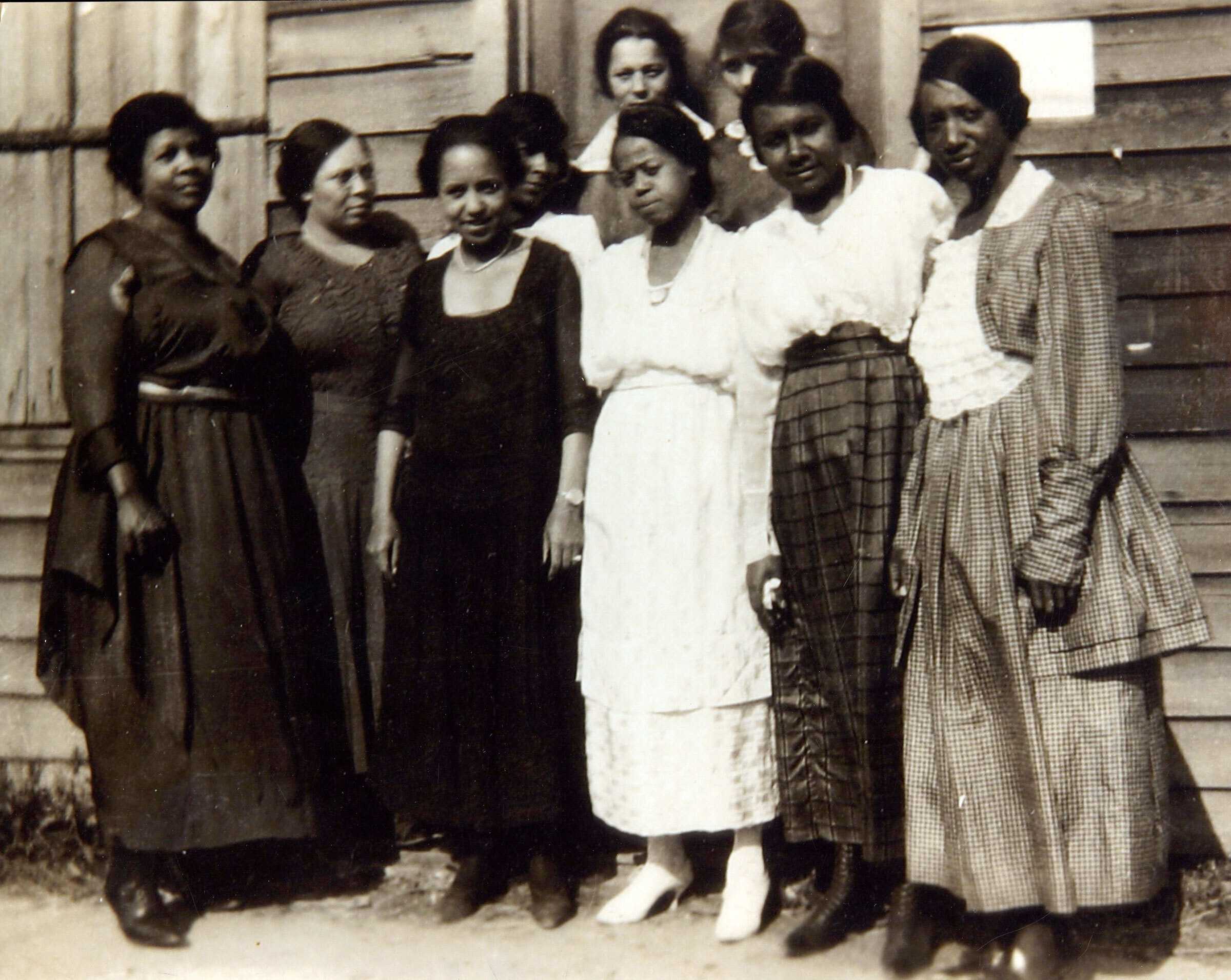 Black and White photograph of a group of African American women standing in front of a wooden building.  All are dressed in long skirts or dresses.