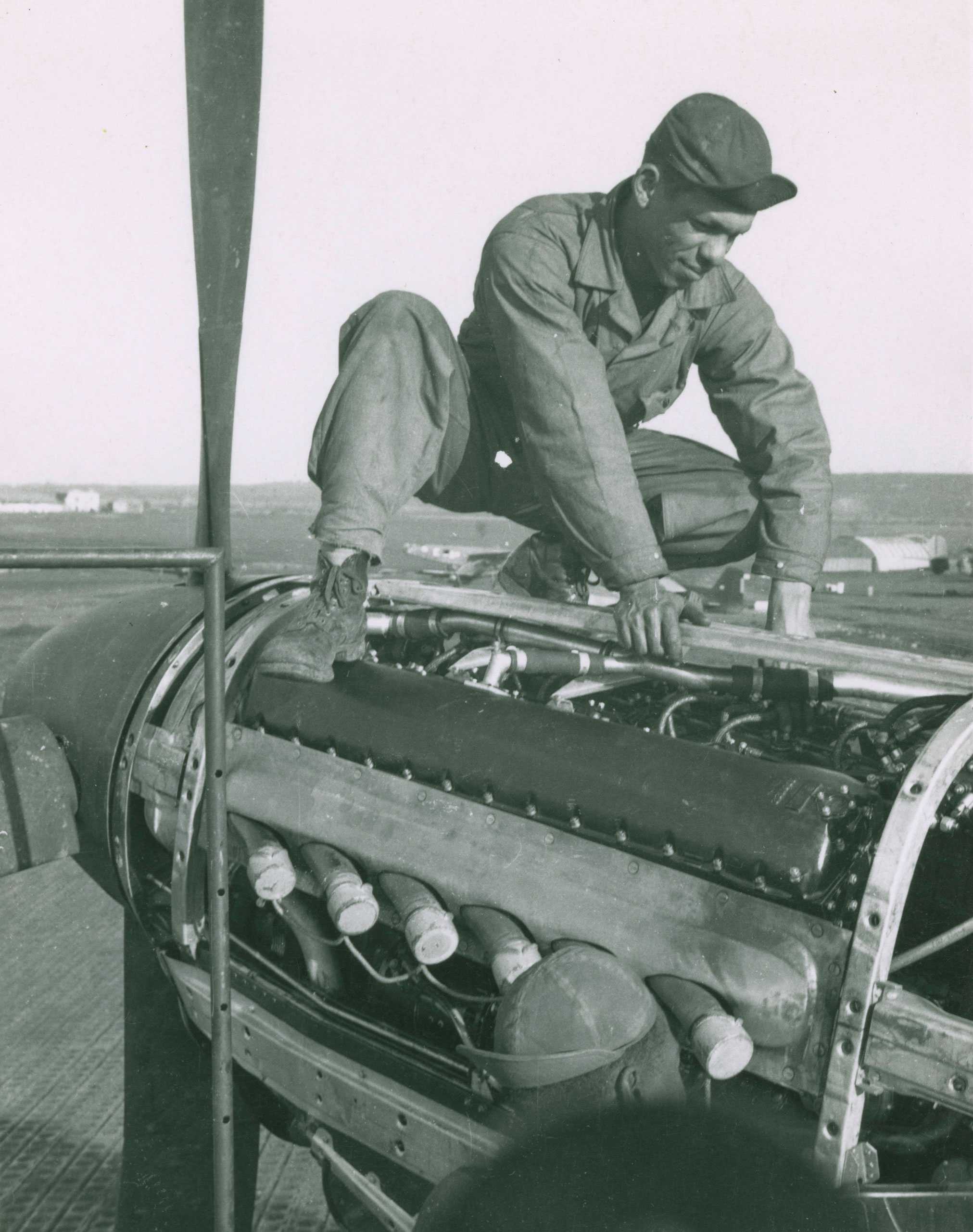 A black and white photo of a Tuskegee airman servicing a P-51 Mustang engine, with another airman nearby.