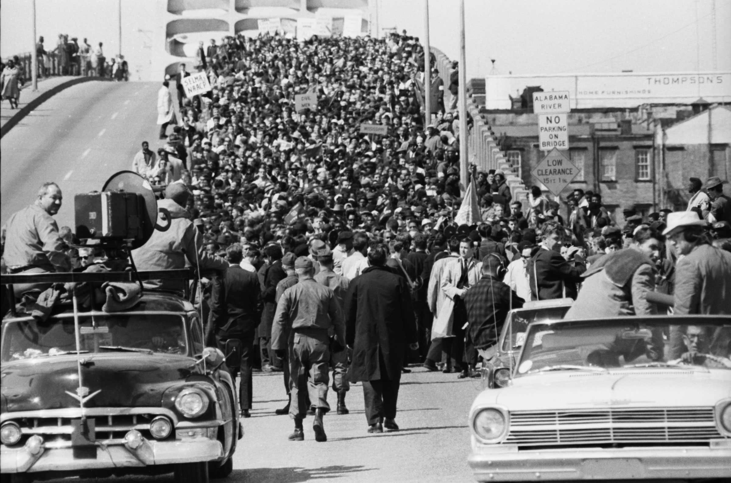 Black-and-white image of marchers on the Edmund Pettus Bridge, with signs held high. Cars frame the bottom, and military personnel approach the marchers.