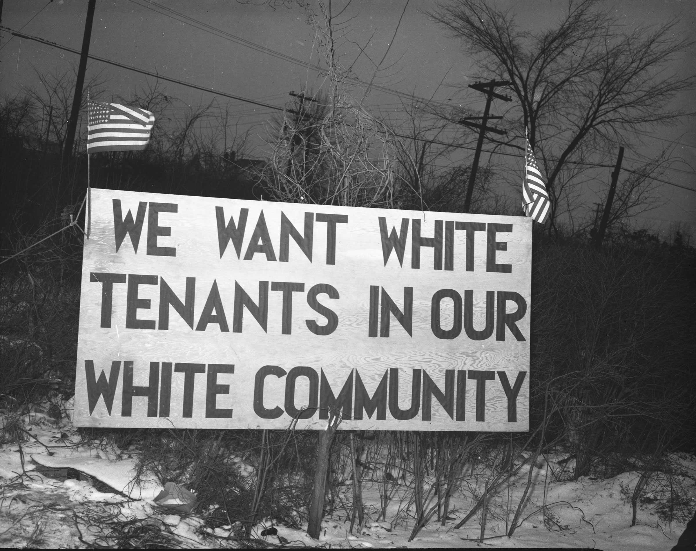 Black and white photograph of large sign in a snowy wooded area.  The sign says "WE WANT WHITE TENANTS IN OUR WHITE COMMUNITY." The sign has two american flags on either corner of the sign.