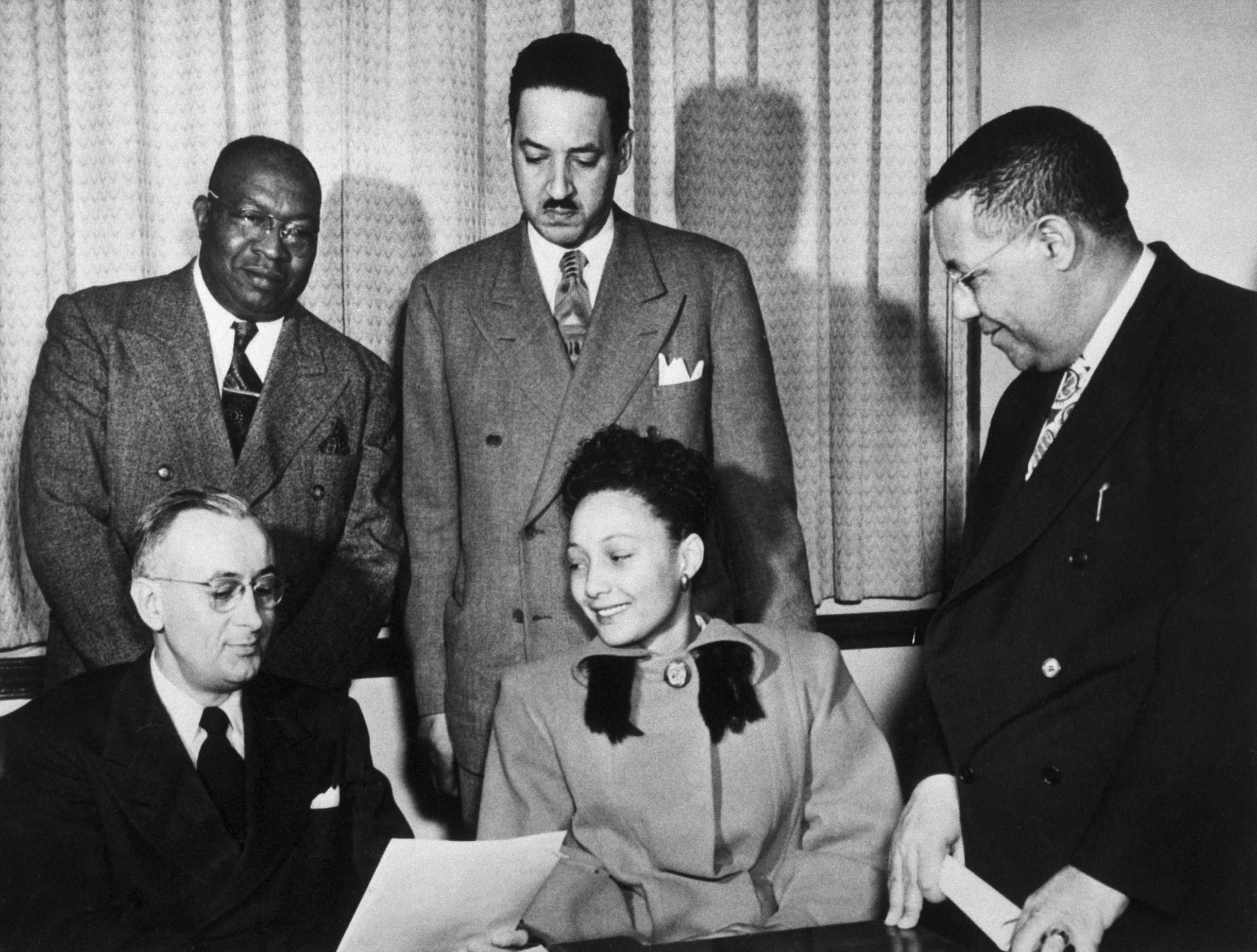 Black and white photograph of woman seated with three men standing behind her and one next to her holding a paper.