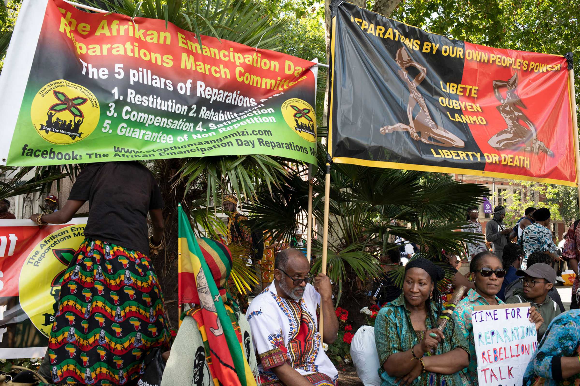 People hold banners and signs demanding reparations. Many of the flags and banner use black, green, and red colors.