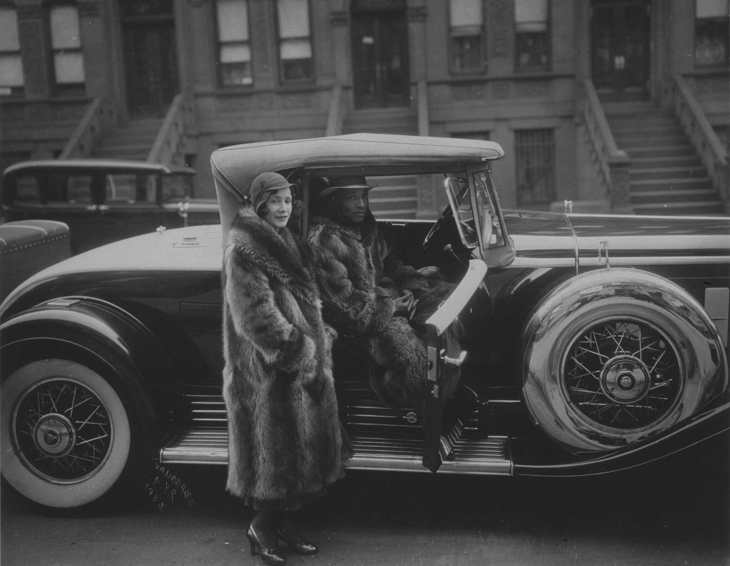 Black and white photograph of a man and women dressed in full length racoon coats.  The man is seated in a car with door open with woman standing next to the car.