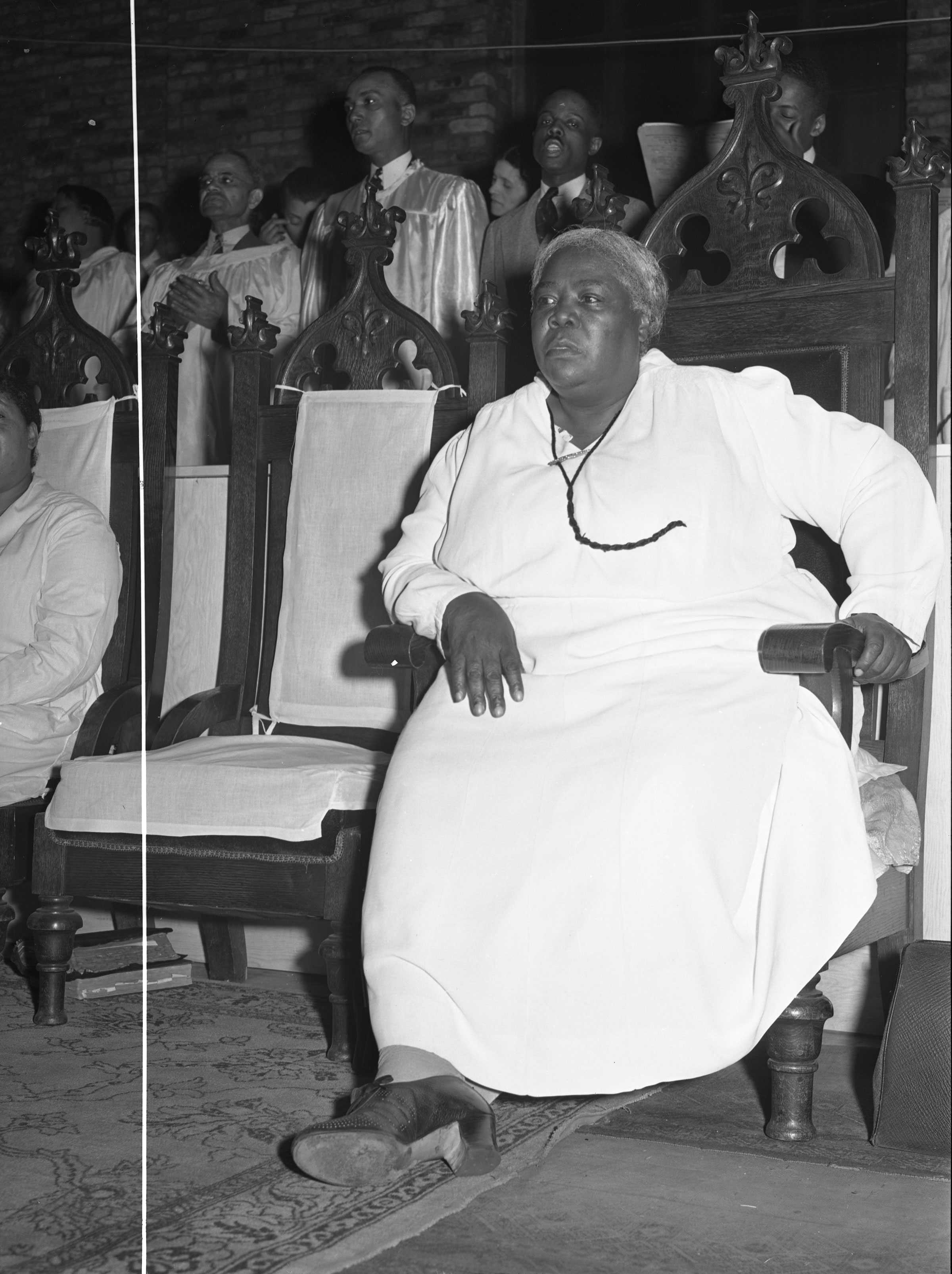 Black and white photograph of Black woman seated in a decorative wooden chair.  She has grey hair pulled back and is dressed in a white dress.