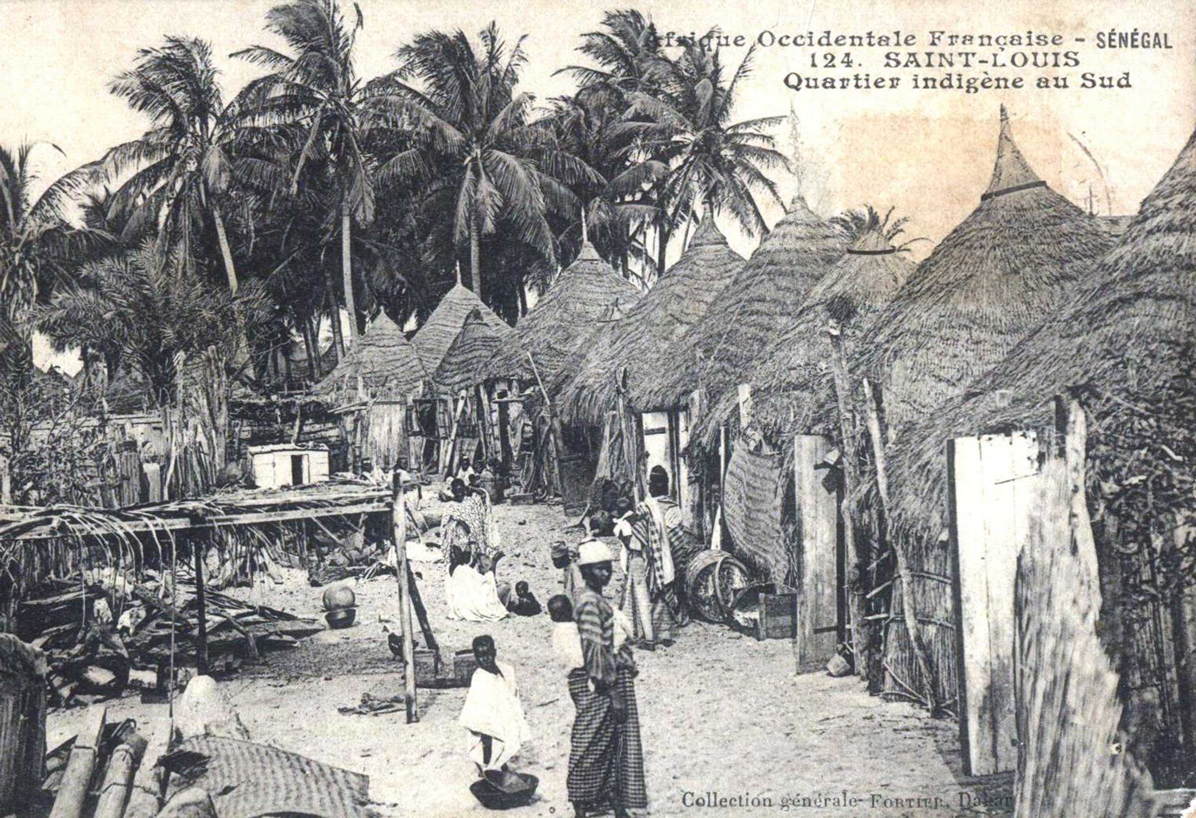 A black and white photograph of a village in Senegal. Grass huts are lined up next to each other.