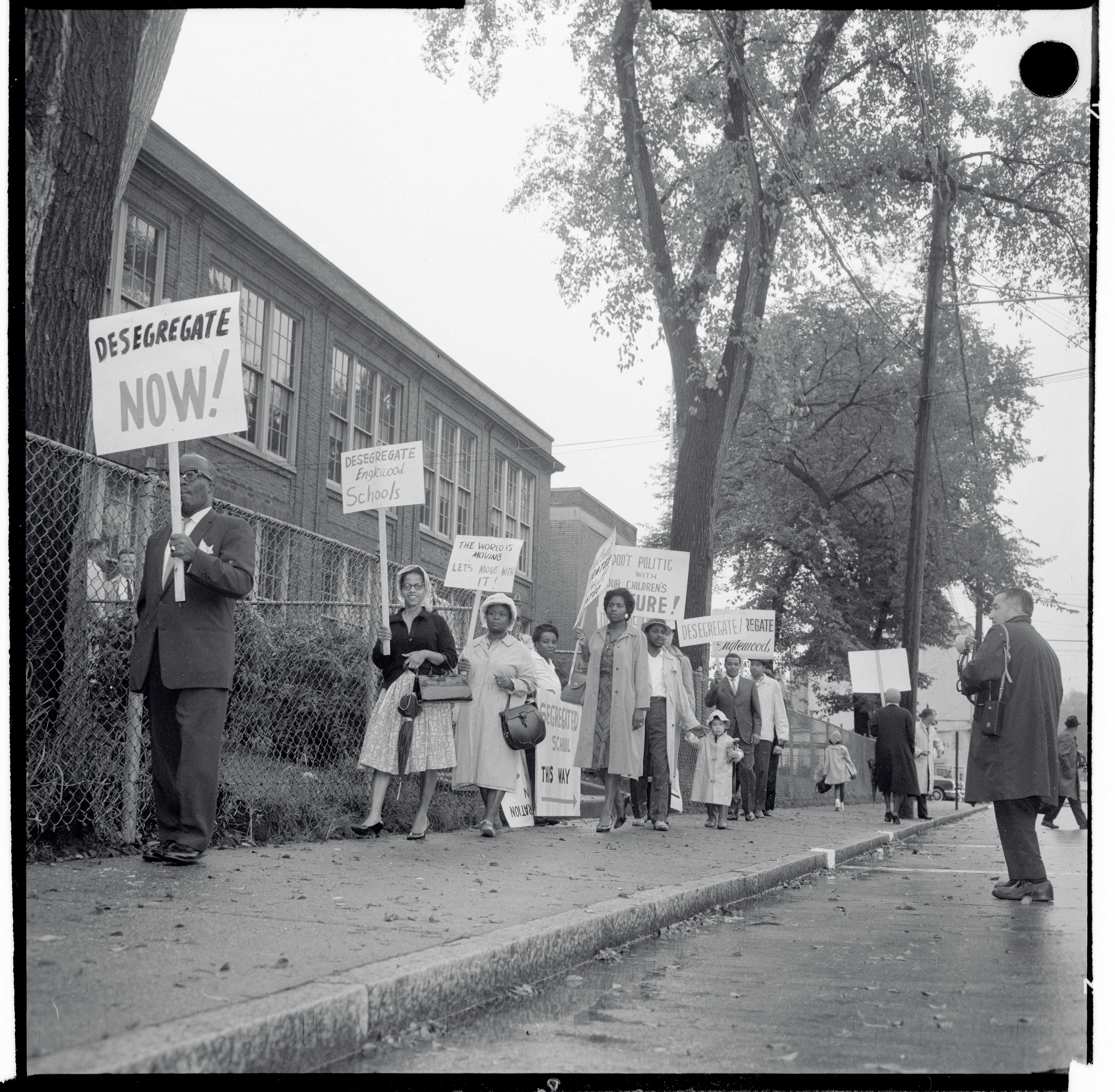 Black and white photograph of people protesting holding signs that say "Desegregate Now".