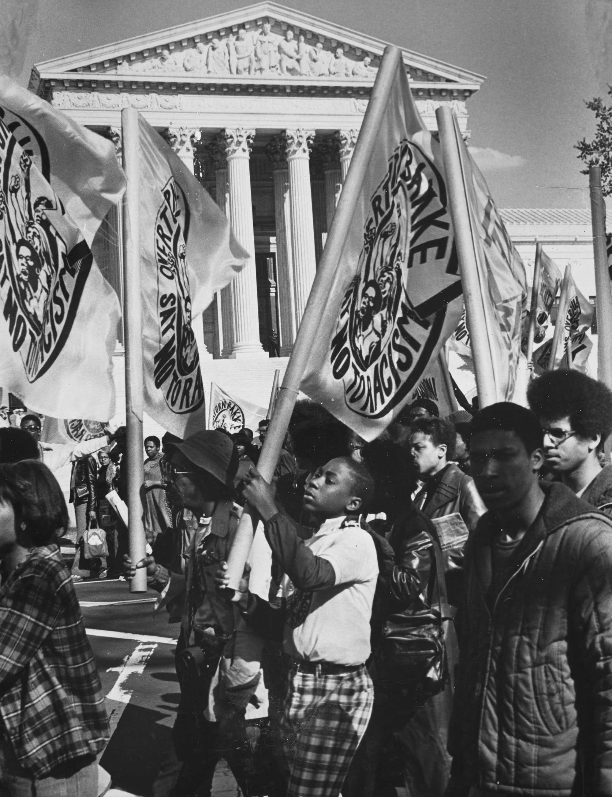 A black-and-white photograph of young adults and adults marching with flags that say "Overturn Bakke Say No to Racism" in front of the Supreme Court.