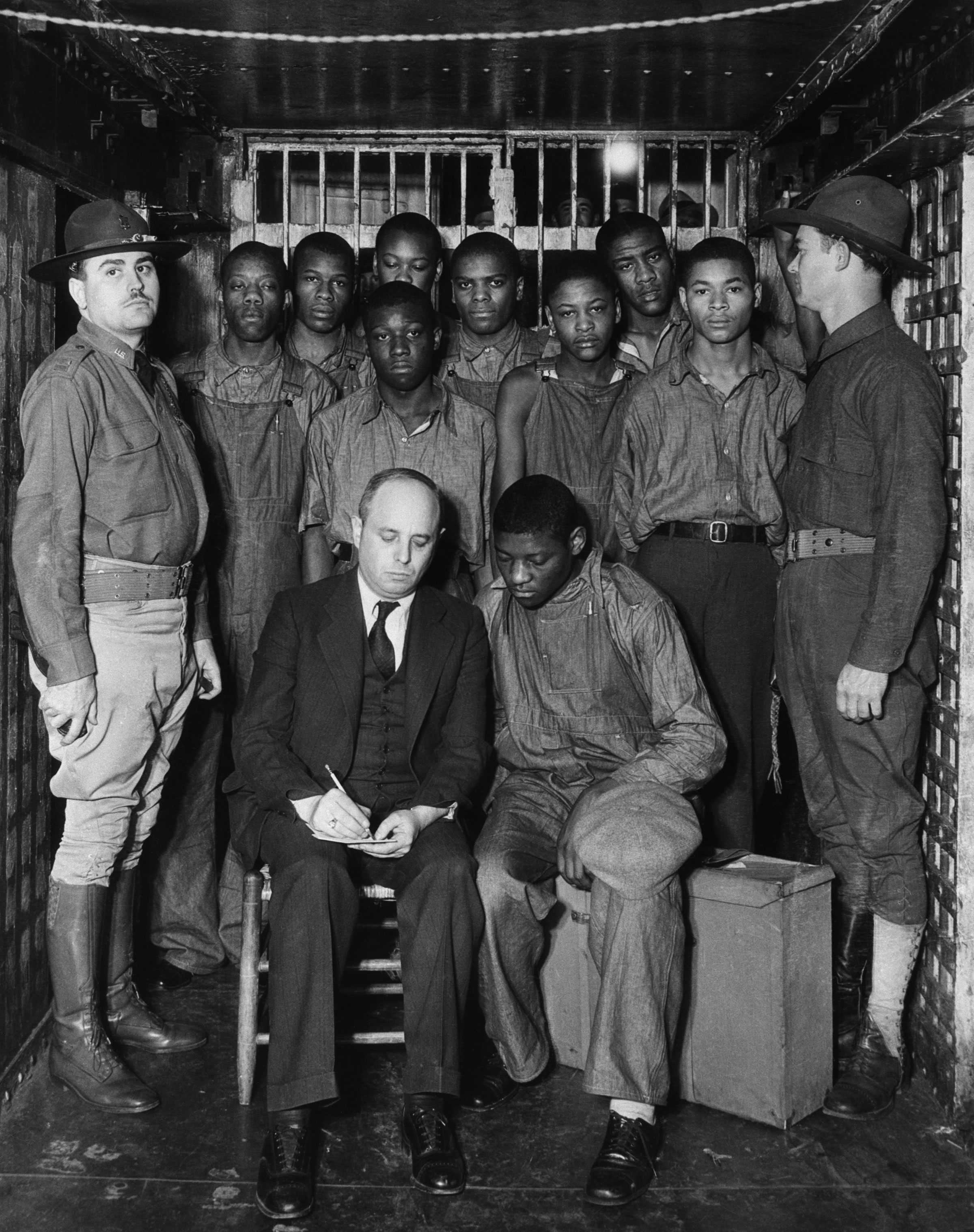 Black and white photograph African American men standing in a prison cell as a seated white man writes down notes.  There is another seated African American man seated next to him.  Two white guards stand on either side of the group.