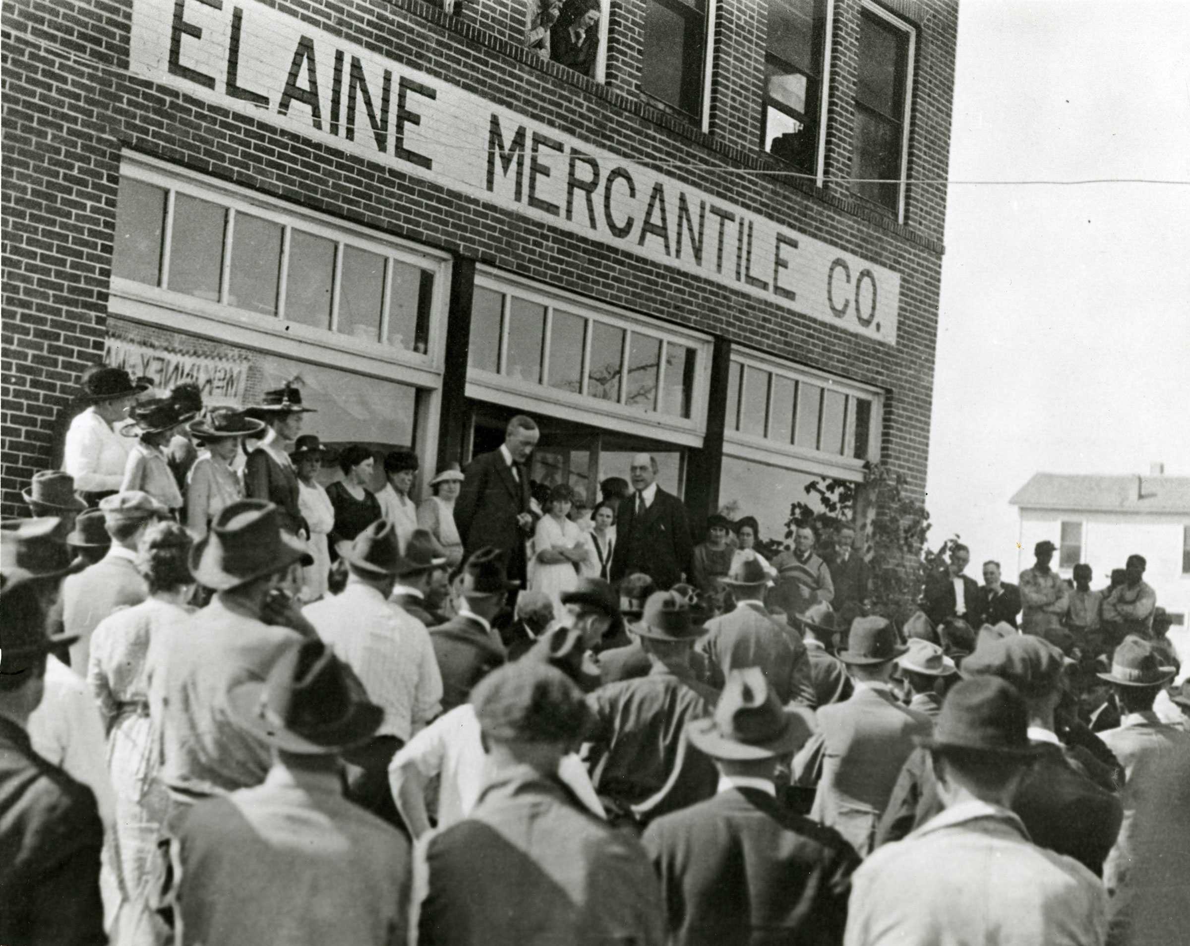 Black and white photograph of large  group standing in front of building identified as "Elaine Mercantile Co." There are two men dressed in suits addressing a large crowd.