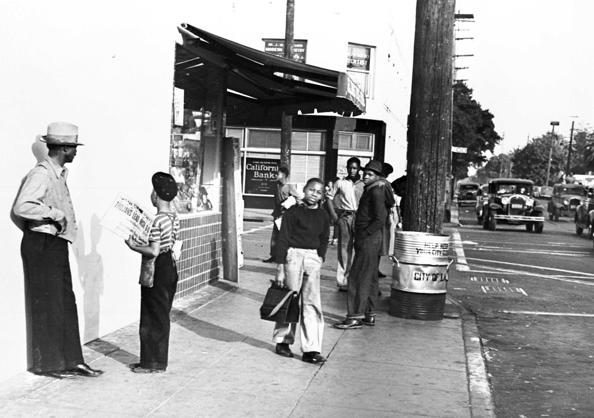 Black and white photograph of street corner in Los Angeles, California.  A man talks with a newsboy and others stand in the distance.