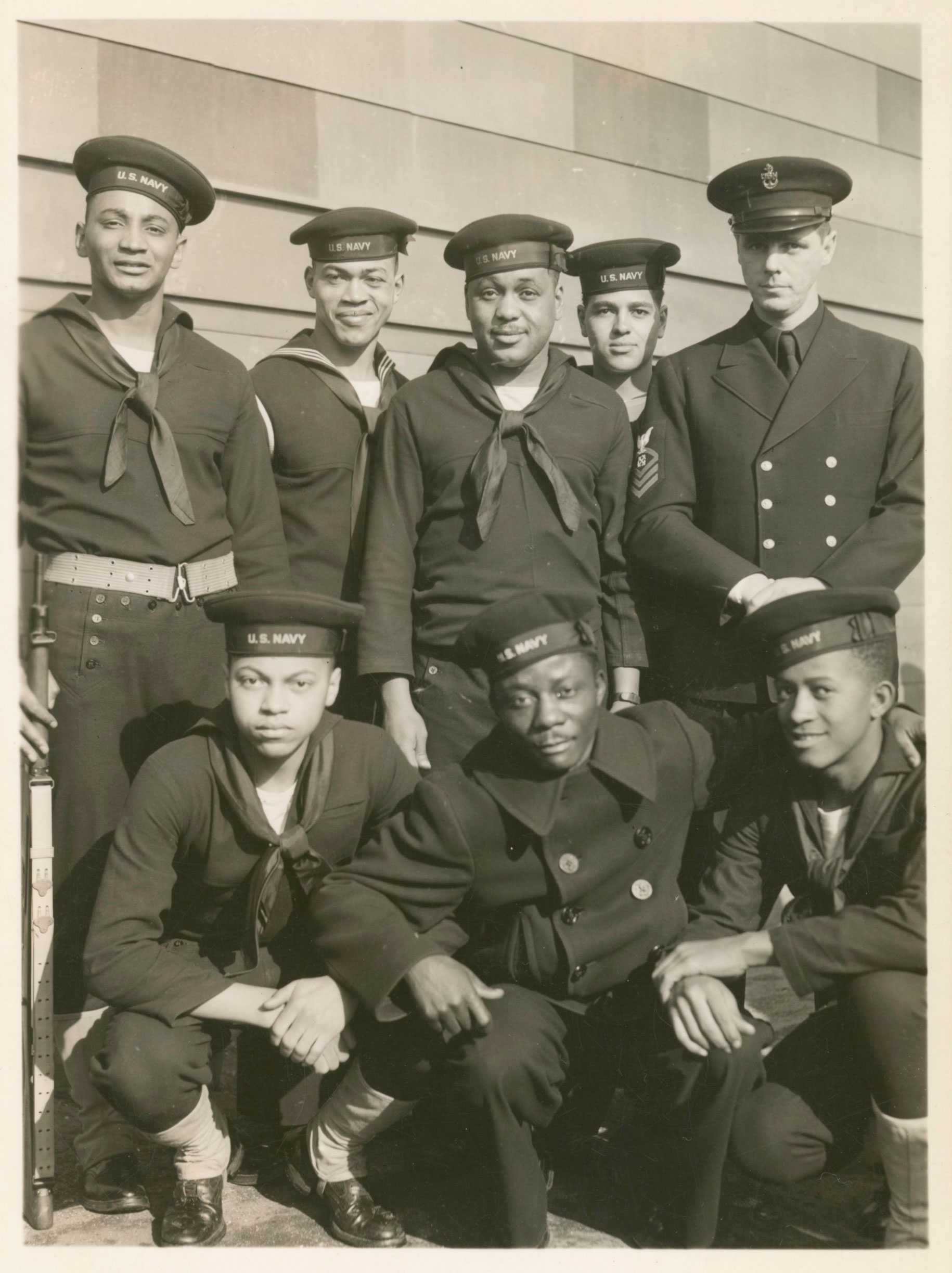 A black-and-white photographic postcard of eight members of the U.S. Navy. Five men stand behind three men who are crouching.