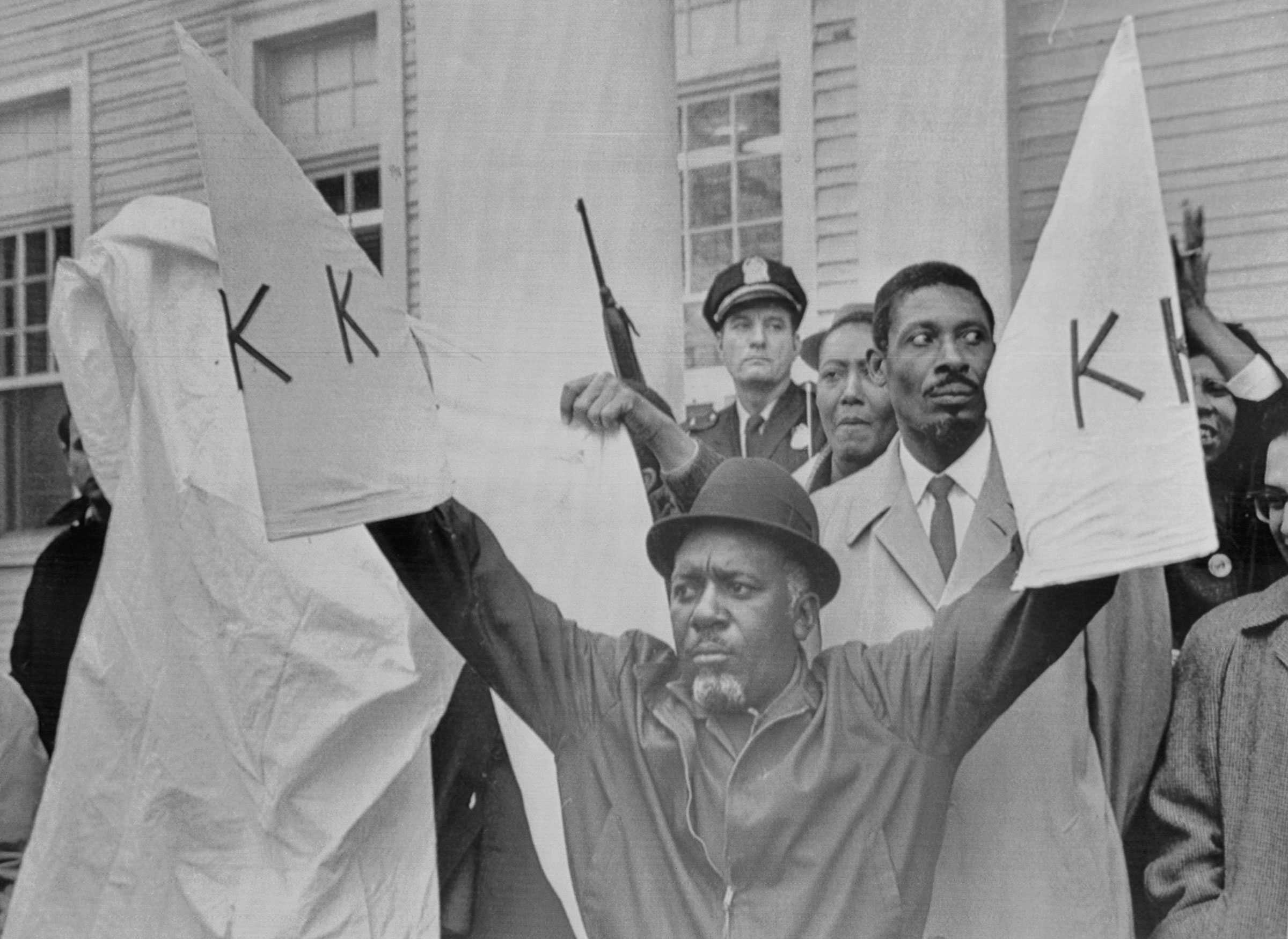 Charles Sim holds up replicas of Ku Klux Klan robes in front of a crowd. He looks determined as he looks into the crowd.