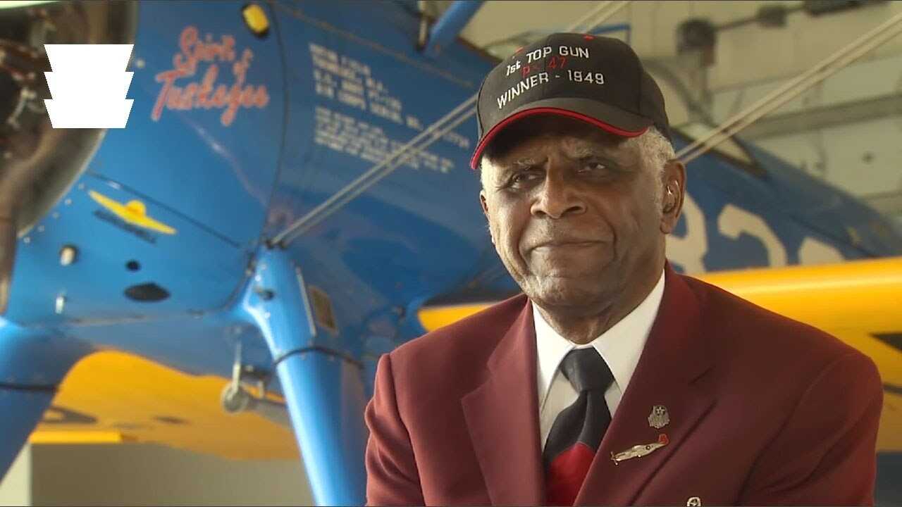 Older man in an airforce hat sits for an interview infront of an plane.