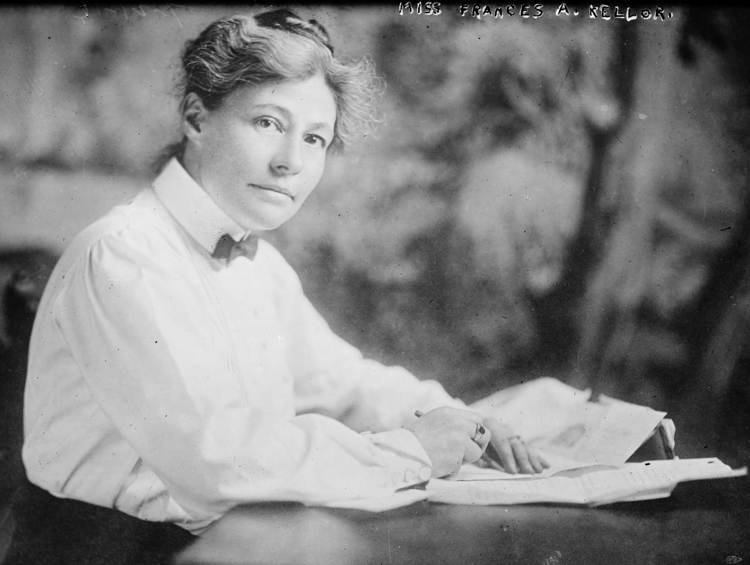 Black and white photograph of young woman seated at desk with pencil and paper on the table in front of her.  The woman wears a white blouse with a bow tie.