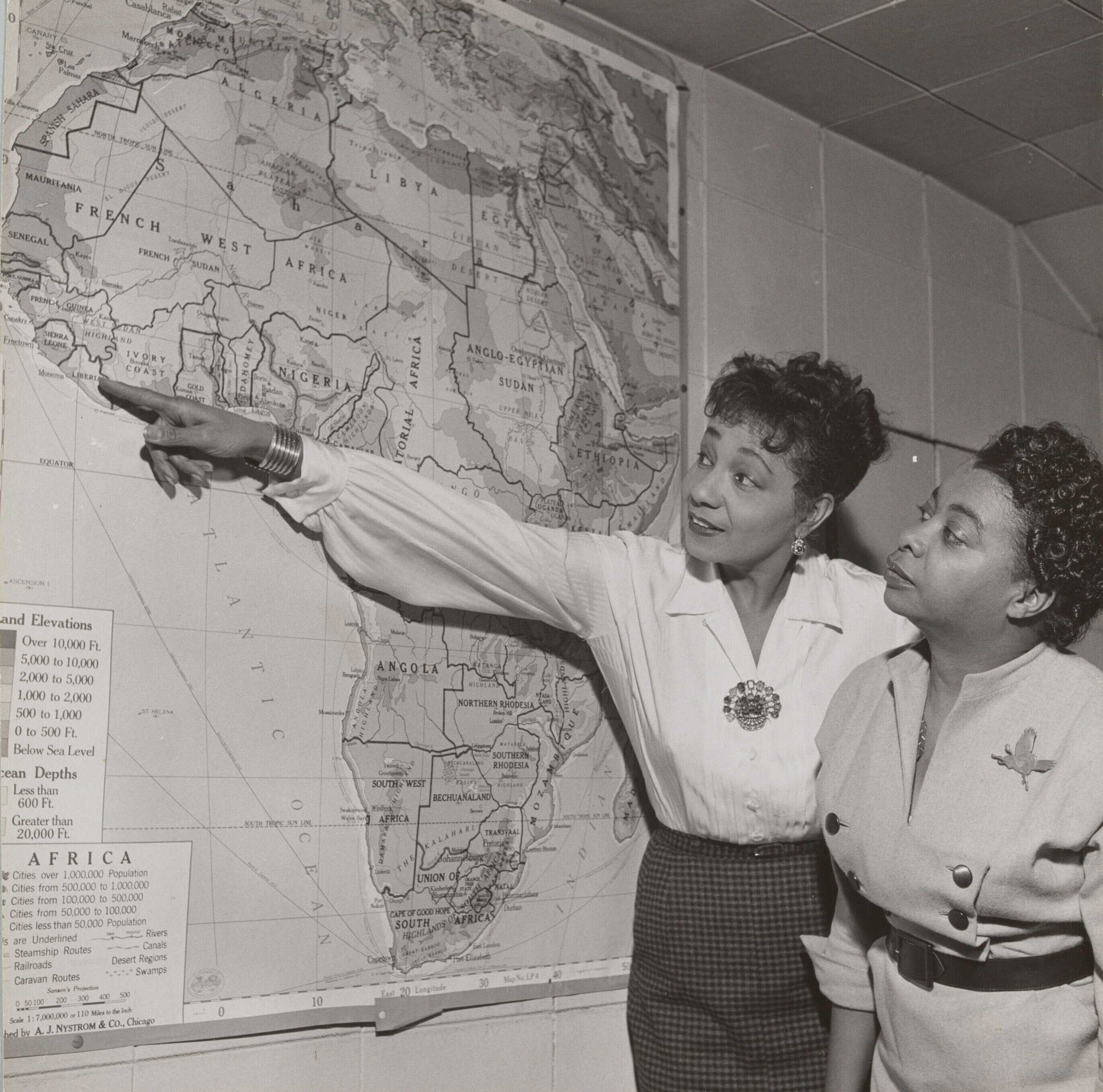 Black and white photograph of Etta Moten Barnett and another woman examining a large map of Africa hanging on the wall. Wearing a long sleeved white blouse with a brooch at the neckline and a grey straight skirt, Etta Moten Barnett stands to the side of the map and points to the country of Liberia with her right arm outstretched. The woman standing next to her wears a light grey dress with double breasted black button up the front and a black belt.