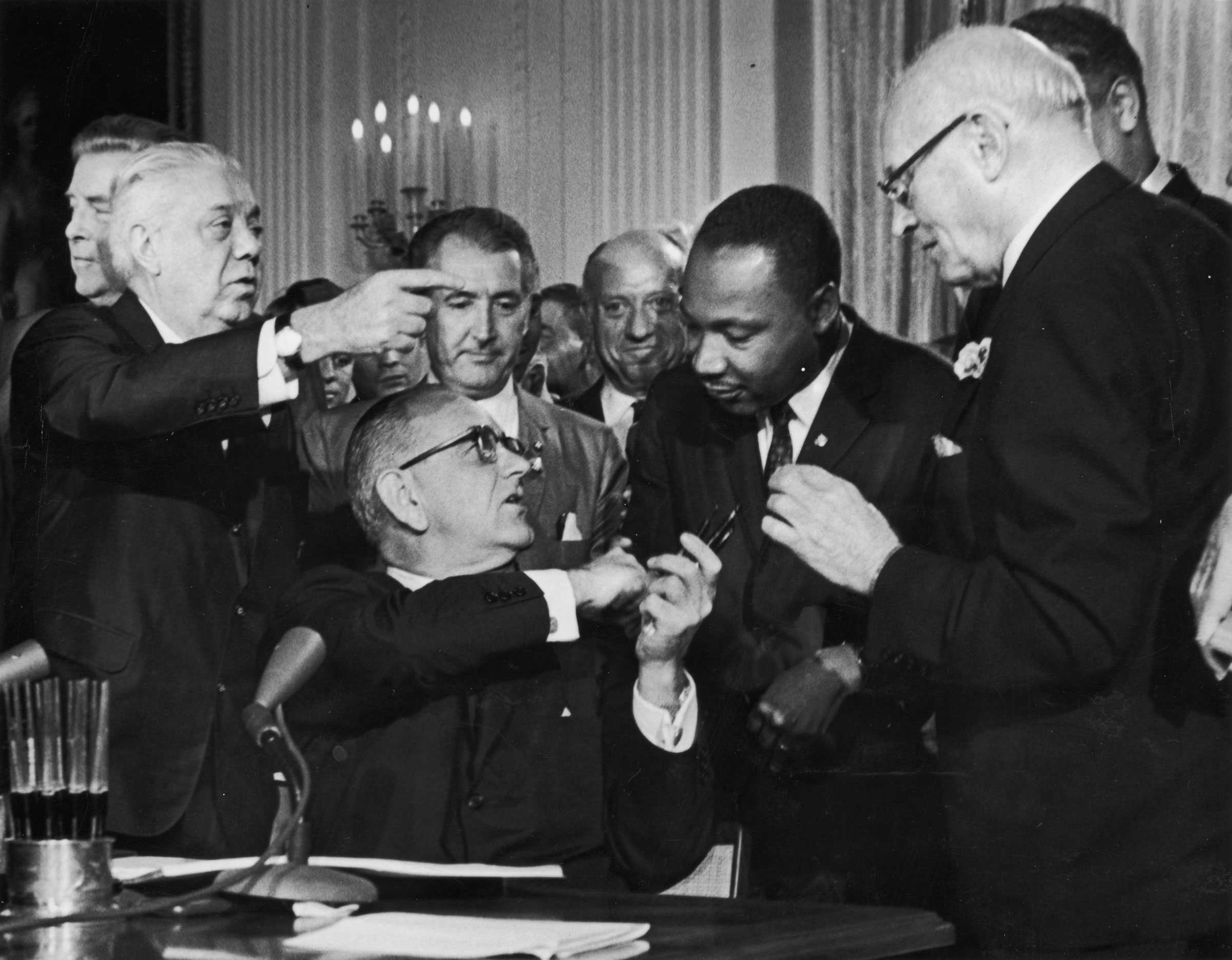 A black and white photo of  President Lyndon B. Johnson shakes the hand of Dr. Martin Luther King Jr. after signing the Civil Rights Act.