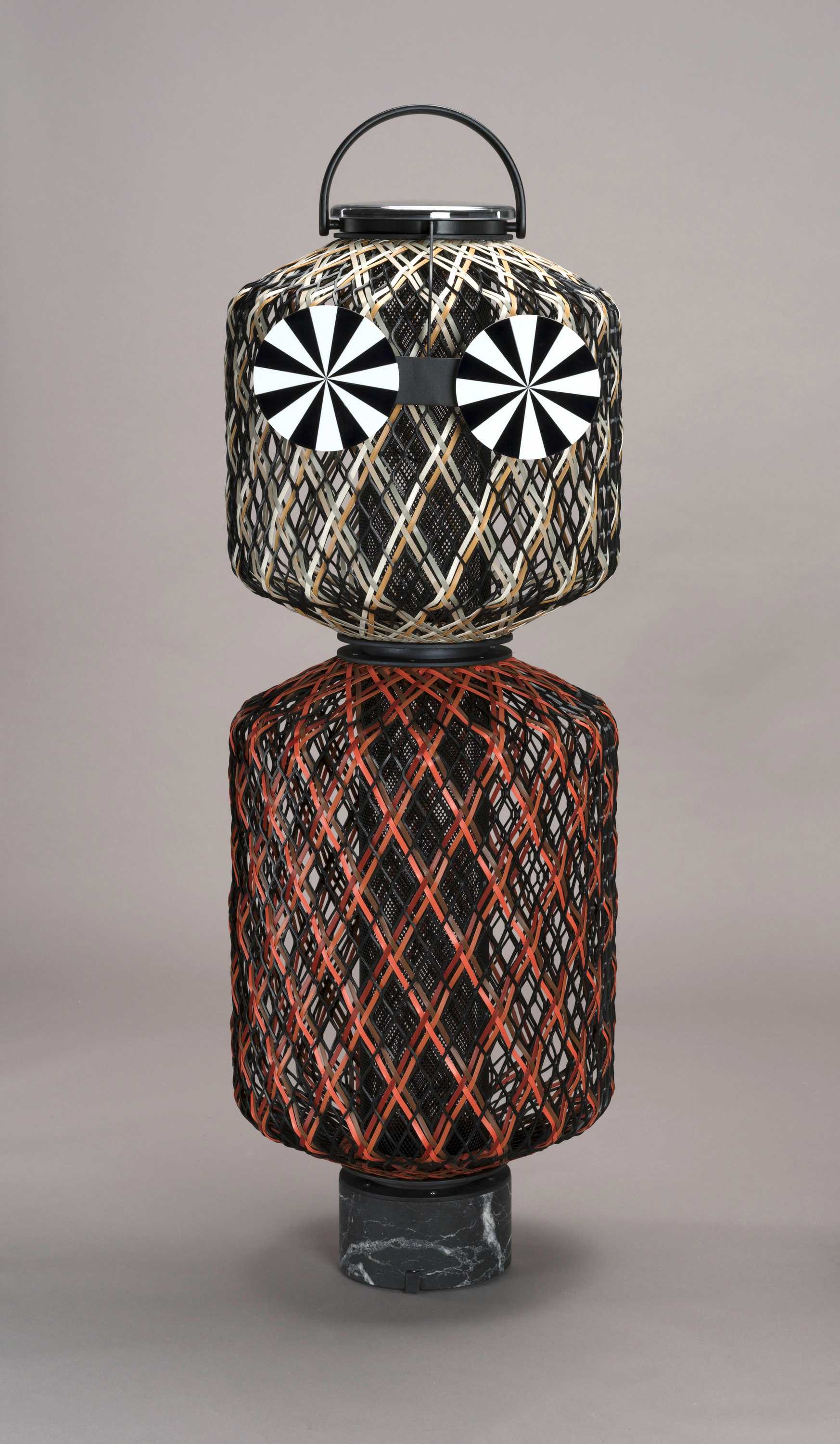 Two stacked hand-woven lantern shades, with the bottom lantern larger than the top. The frame of each lantern is made from black coated aluminum. The top lantern is in the color shade ‘Terra’ with the interwoven fibers in light brown, light grey, or beige. The bottom lantern is in the color shade ‘Rock’ with the red, orange, and brown interwoven fibers.