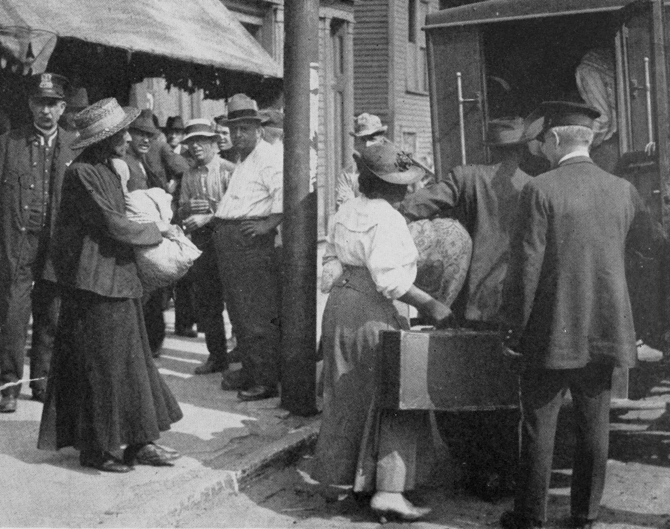 Photograph showing women being loaded into a van.  They are holding suitcases and large bags.  Policemen look on as well as unidentified men.