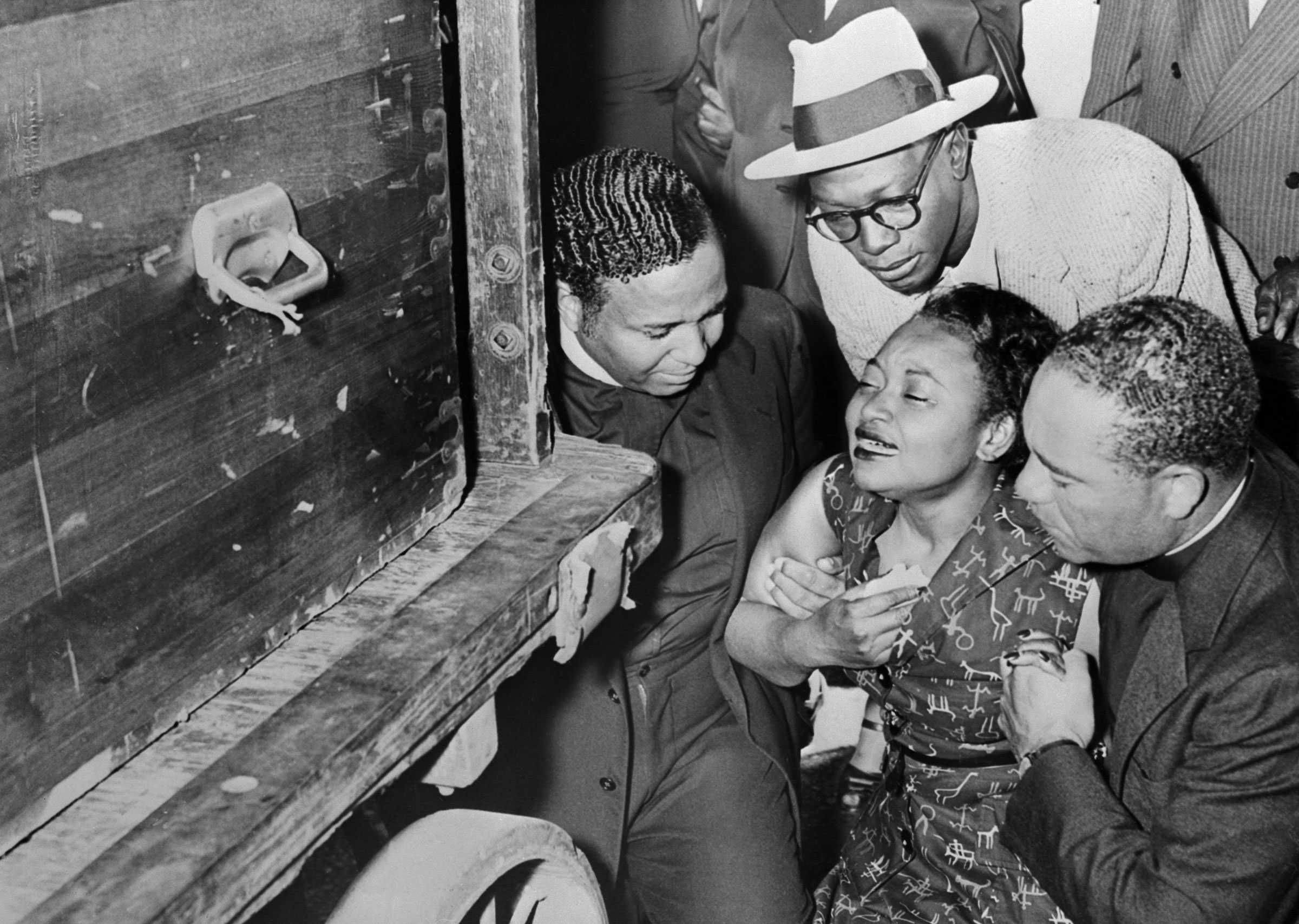 A black and white photograph of Mrs Mamie Bradley weeps aside her son's cassette. She is being held up 3 men, on her side.