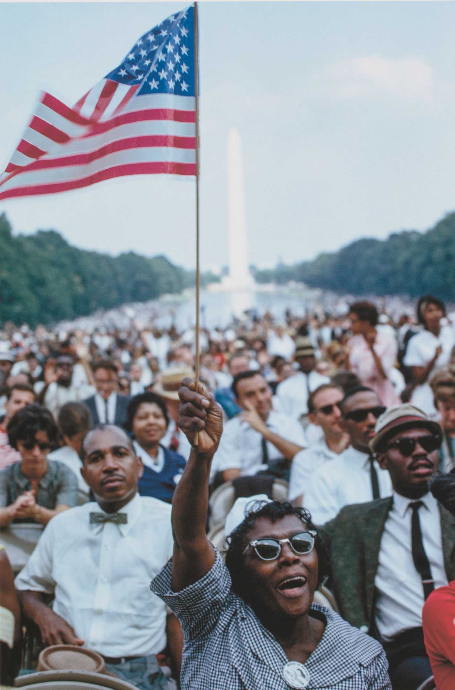 A color photograph of the 1963 March on Washington. A woman waves an American flag in a large crowd with the Reflecting Pool and Washington Monument in the background.