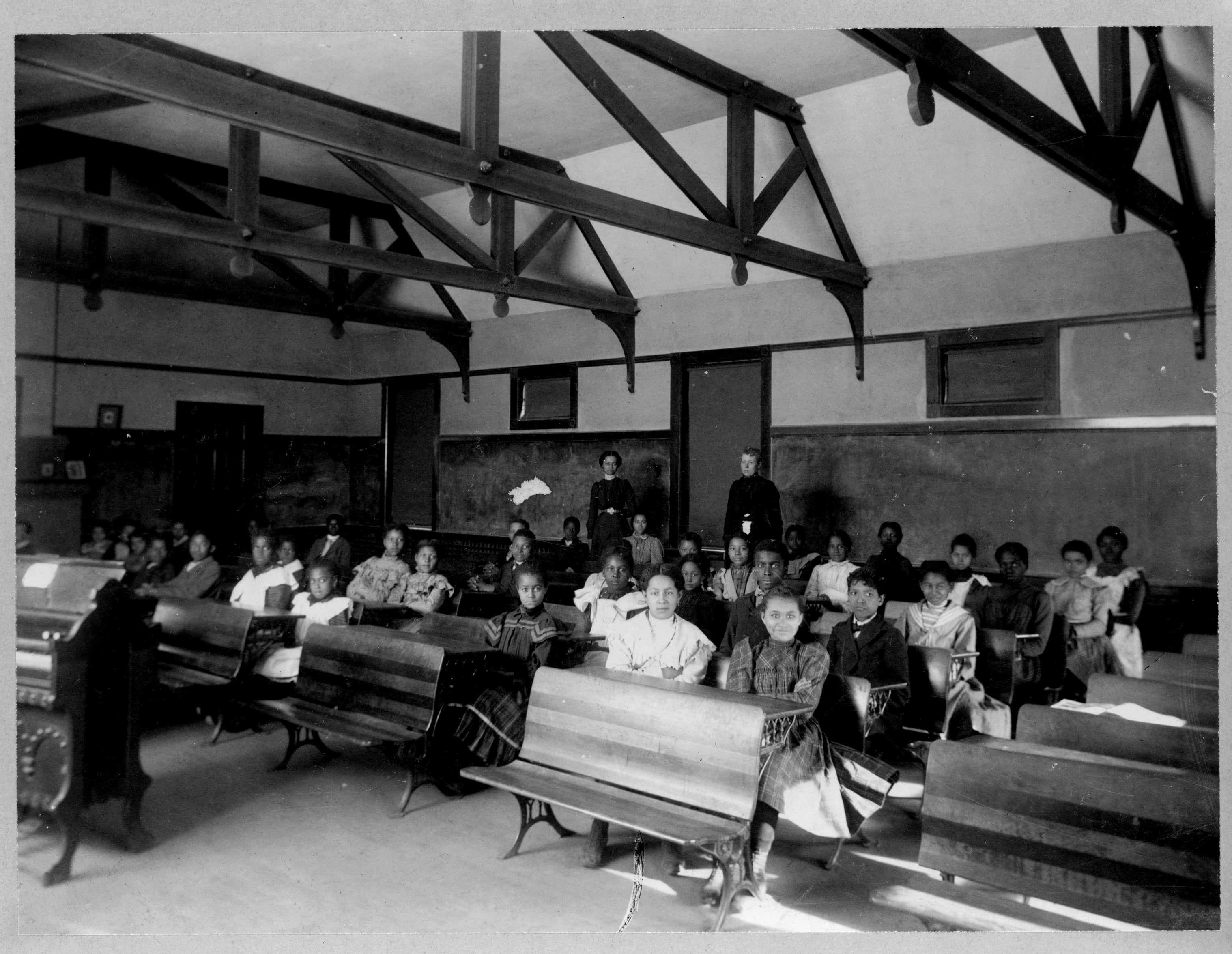 Students inside of a classroom posed for a pictured. Each student is sitting down at desks, while the two teachers stand behind them.