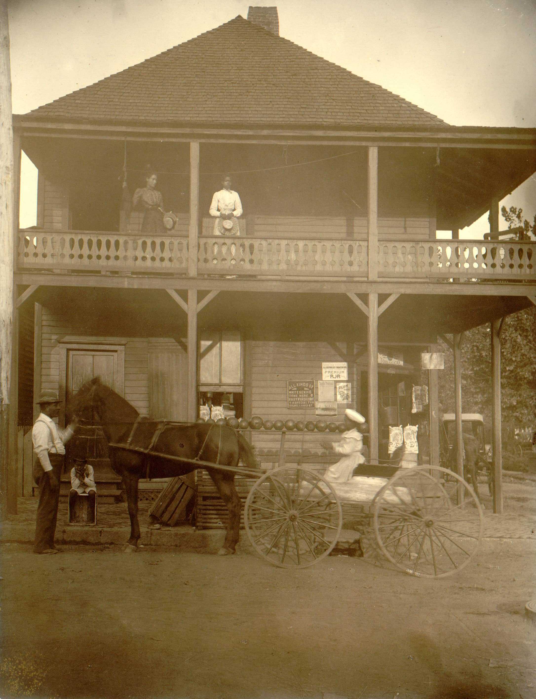 A sepia toned photo of a store. A horse-drawn wagon stands in front. A young woman is on the wagon while a man pets the horse.