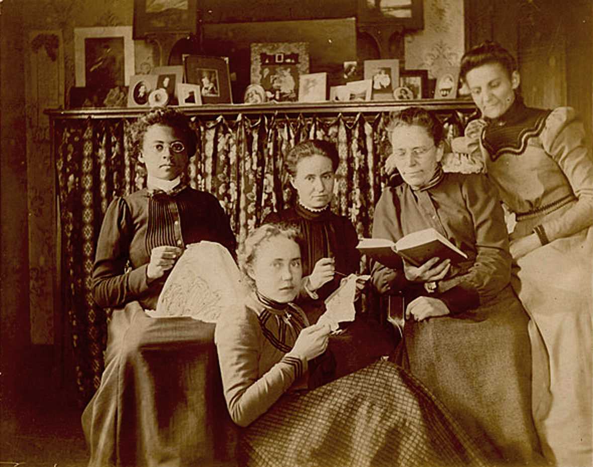 Sepia toned photograph of four seated teachers (one African American and three White) wearing long-sleeved, high-collar dresses reading and doing needlework.