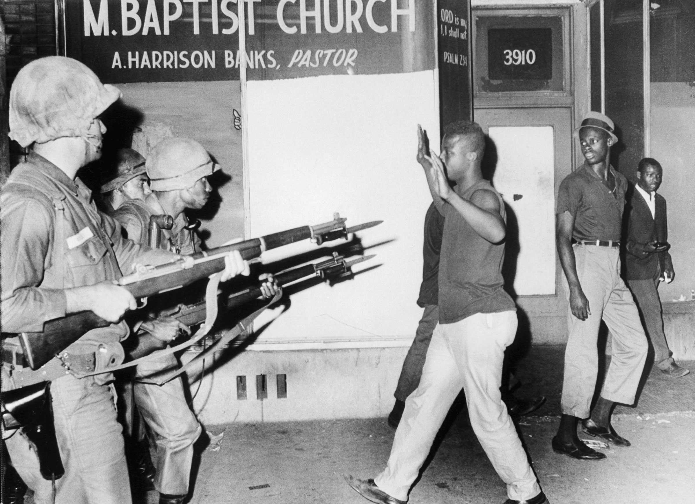 National Guard troops aim rifles with bayonets at a few young black rioters. One reuters walks towards the troops with both hands up.