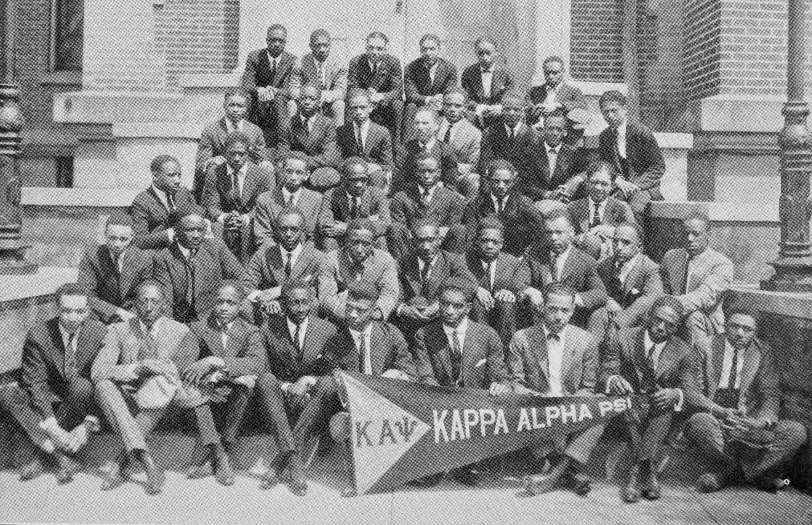 Black and white group portrait of young Black men seated 5 rows deep on the steps of a brick building.  They are all dressed in suits and display a banner that reads "KAPPA ALPHA PSI."