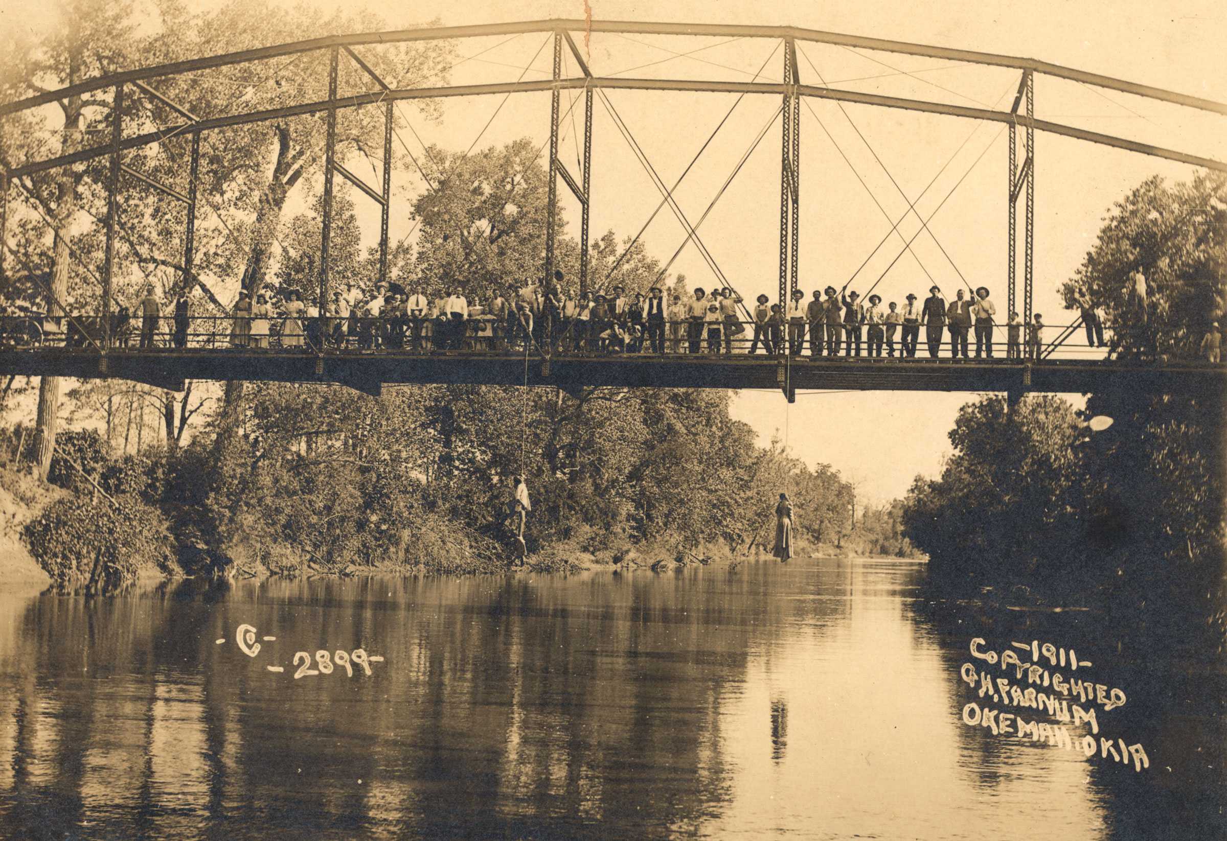 Sepia toned photograph of a crowd standing on a bridge looking at two dangling bodies that were lynched.