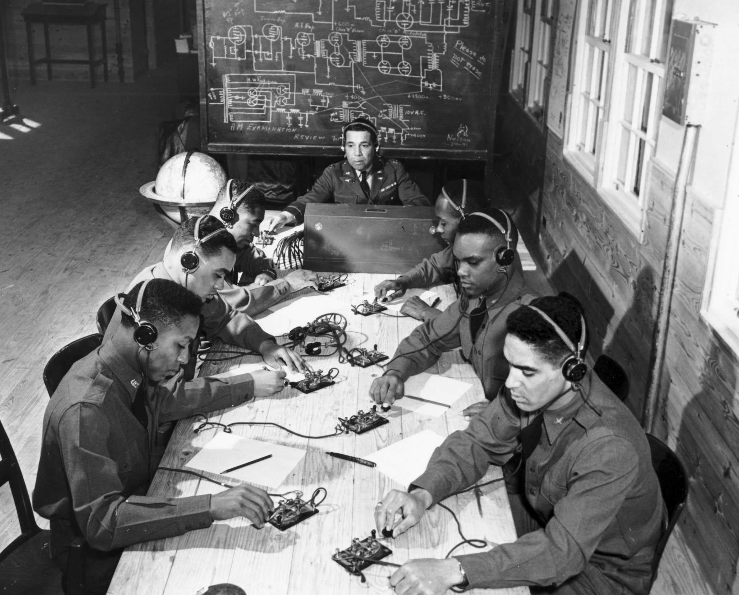 Black and white photograph of African American soldiers wearing headphones seated around a long table while tapping on small device .  They are intensely focused on their work as they listen to direction from their teacher seated at the head of the table.