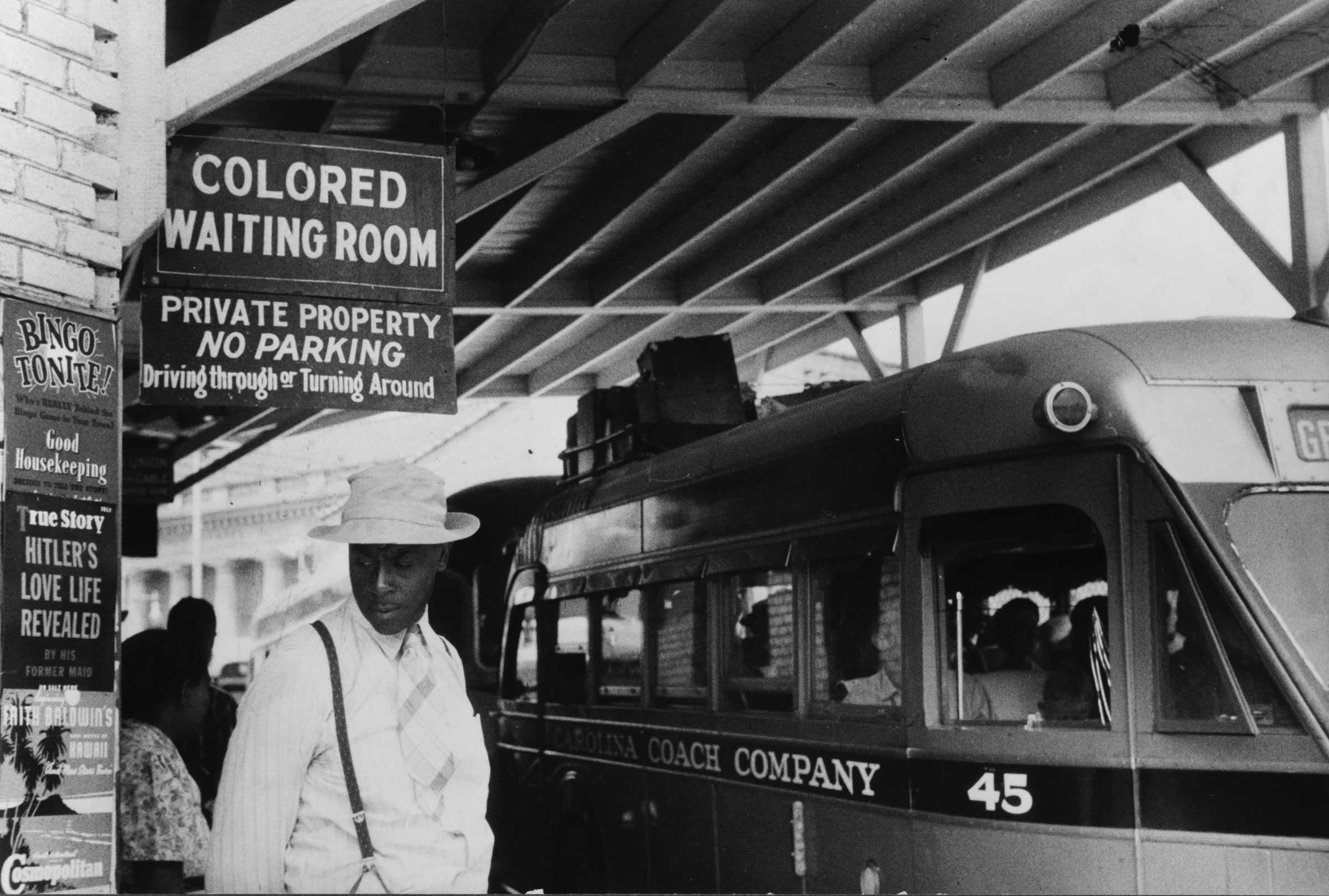 Black and white photograph of exterior of bus station.  There is a black man standing under a sign that reads "COLORED WAITING ROOM" who appears to be waiting for a bus.