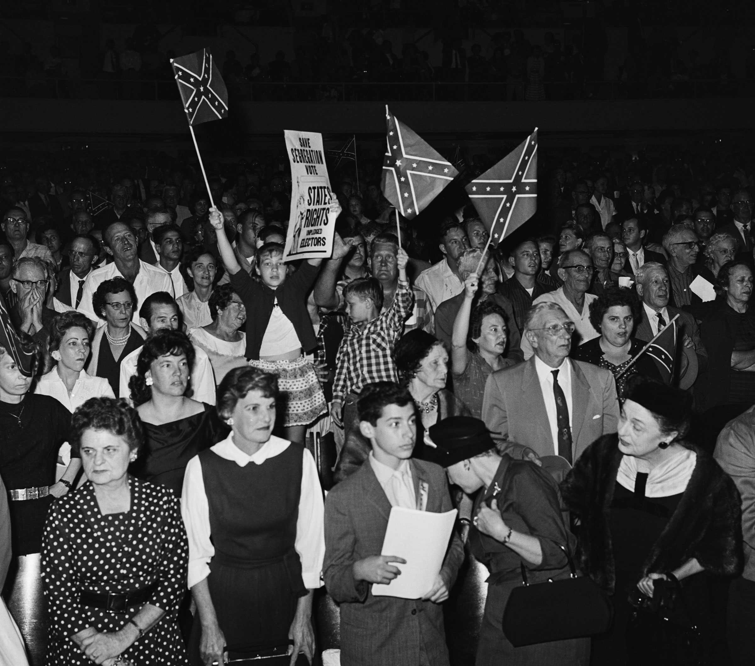 A black and white photograph of a large crowd at a council meeting.Two small children stand on chairs, waving confederate flags, among the adults.