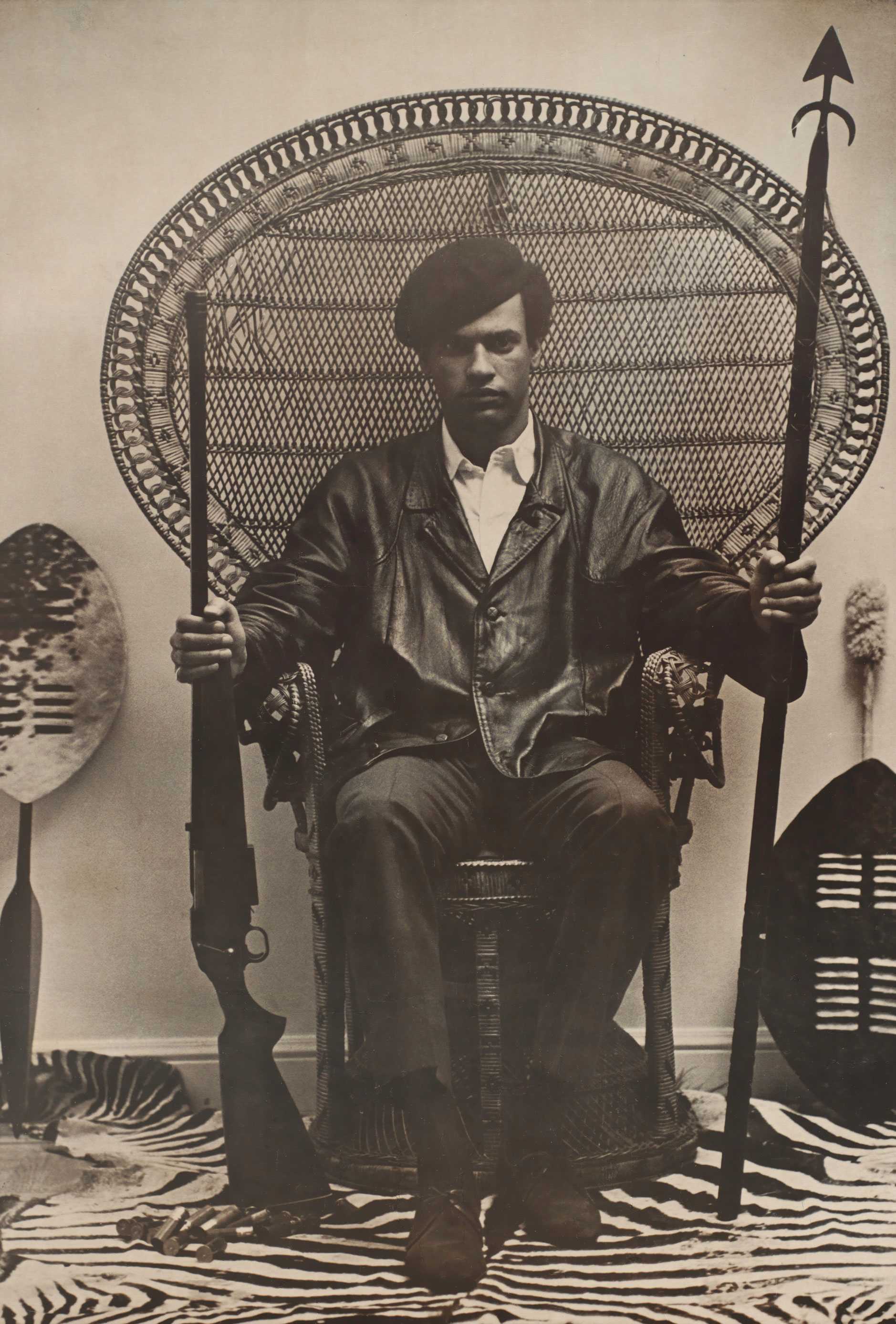 Poster of Huey Newton in a rattan chair, holding a shotgun and spear, with shields and zebra print rug. Text below demands justice against police brutality.