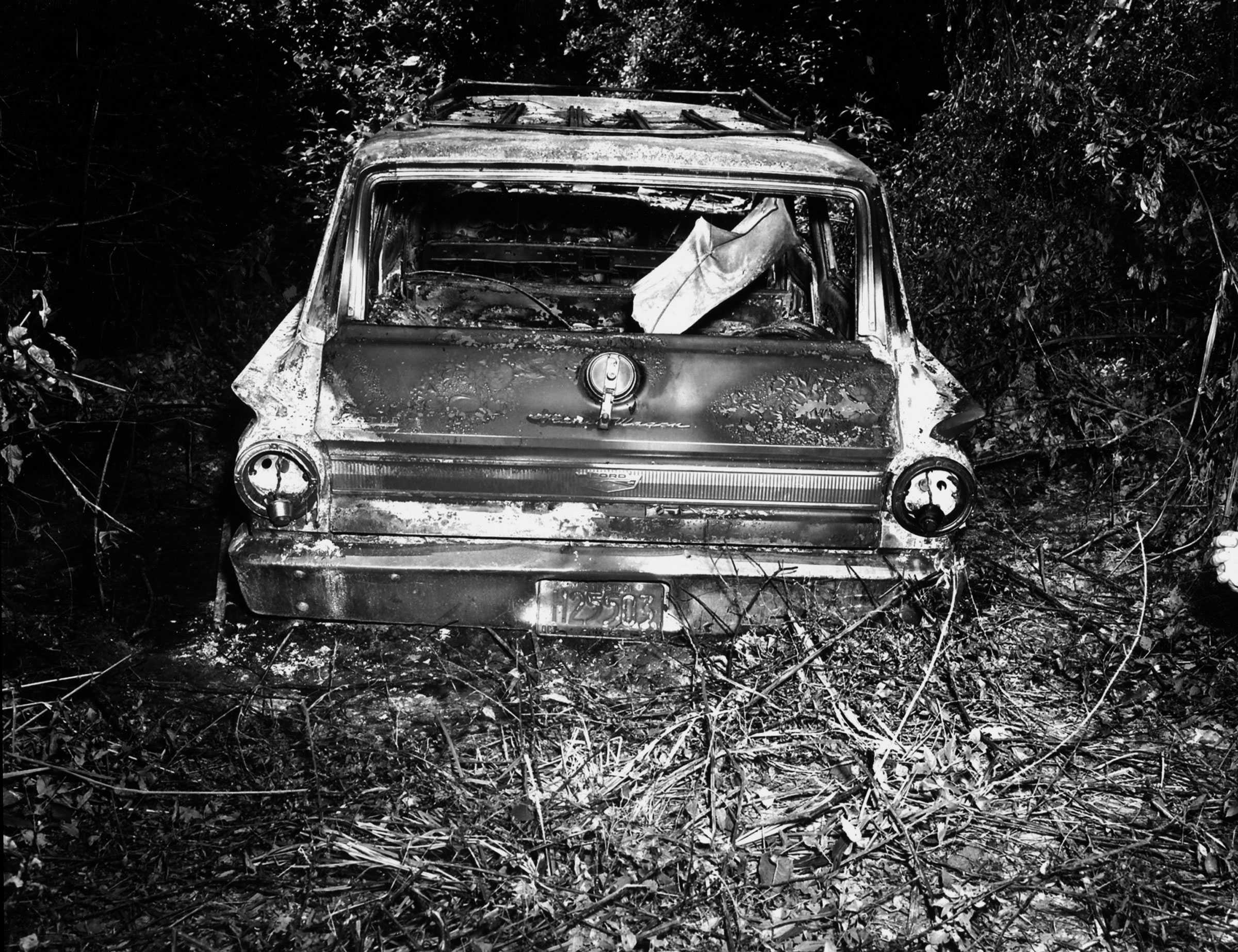 A black and white photograph of a bombed car. The glass of the windows and headlines shattered and the paint has bubbled and peeled.