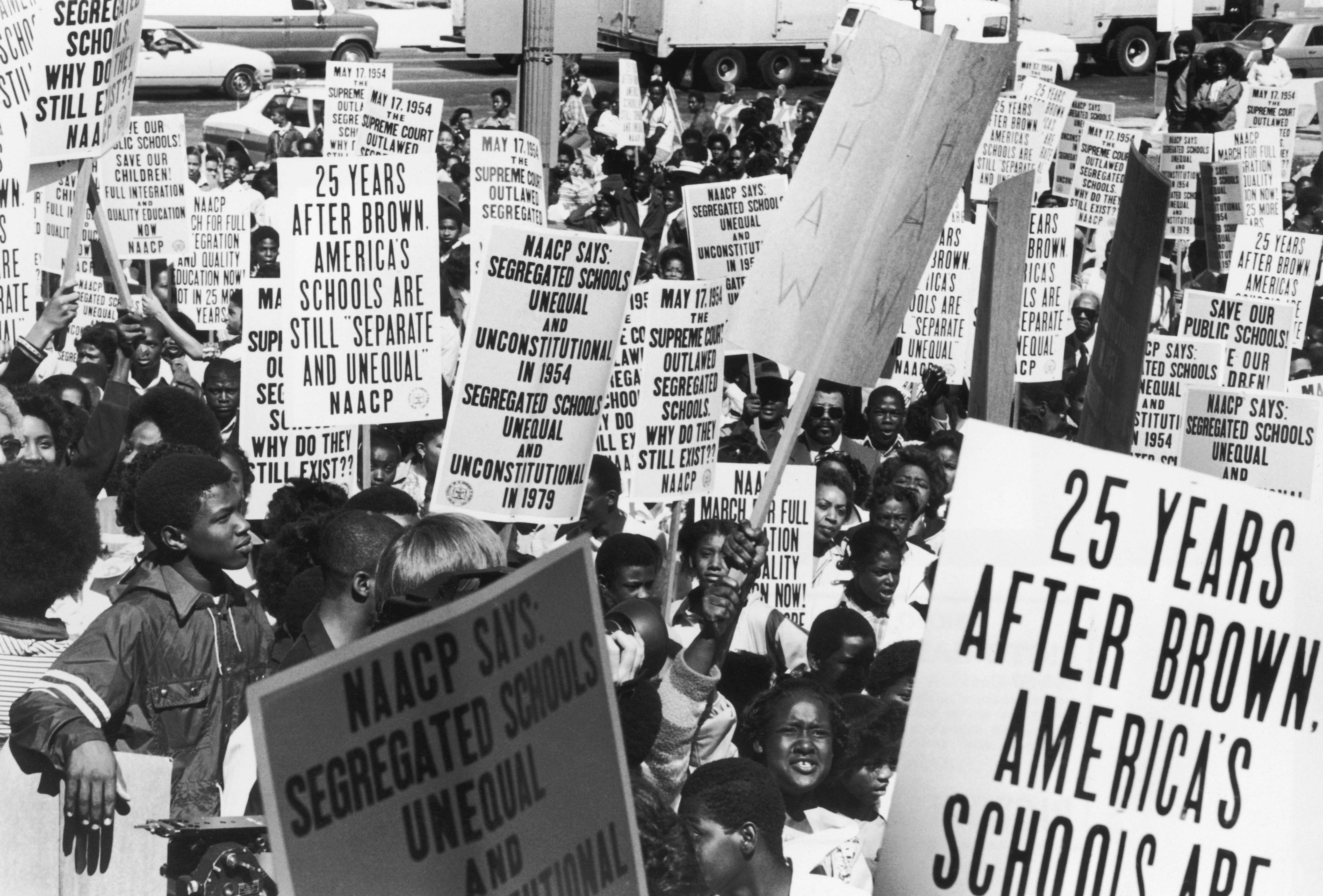 Black and white photograph of 25th anniversary of Brown v Board.  People hold signs saying that America's schools are still segregated in 1979.