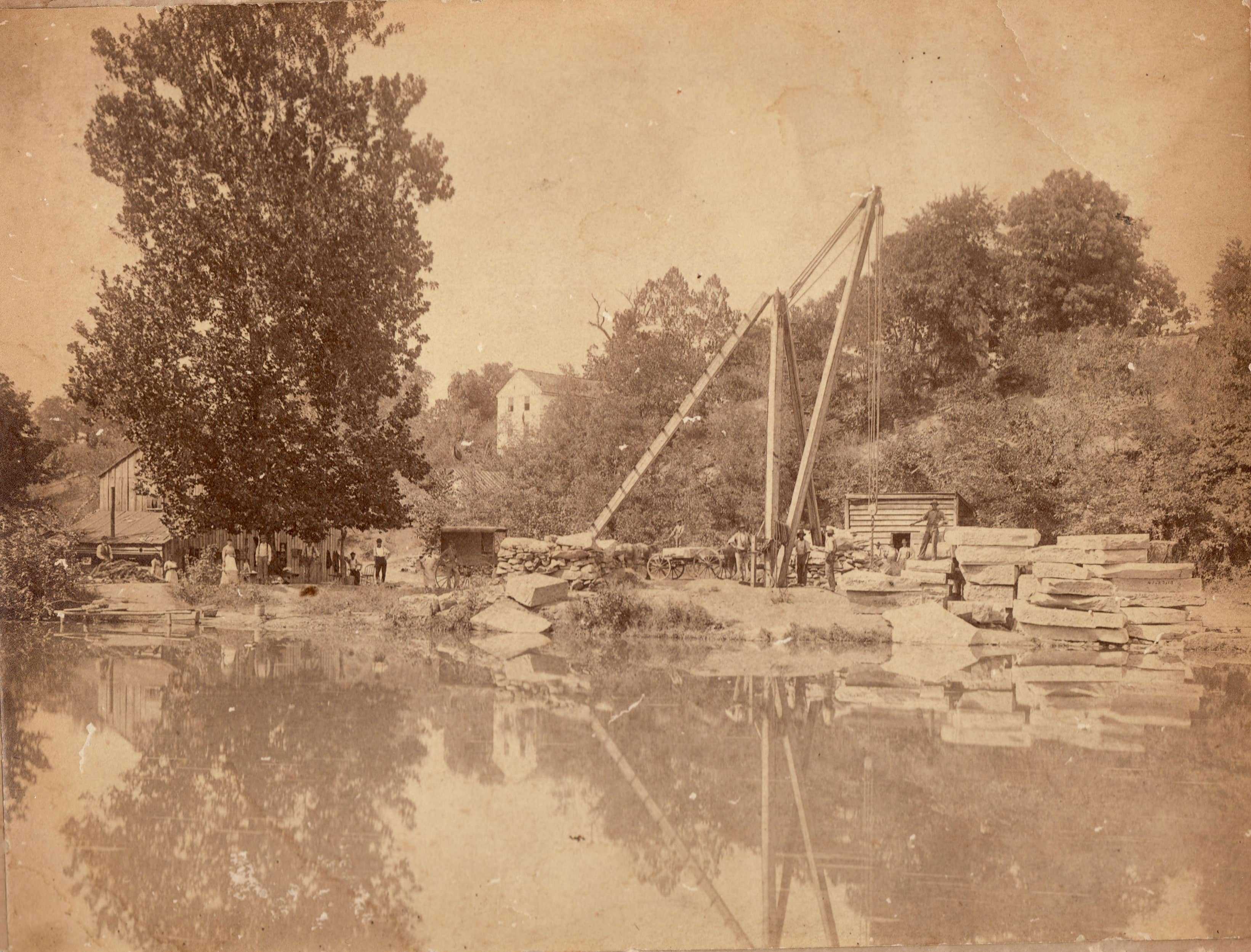 A sepia tone photo of a quarry. People stand along the shore. There is a crane and wagons.